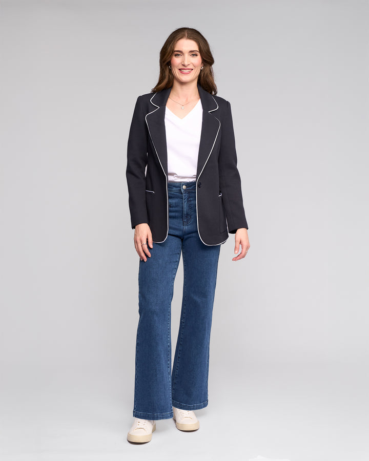 A woman stands and smiles, wearing the Vassalli Ora Blazer - Ink, a white V-neck top, blue flared jeans, and white sneakers against a plain light gray background.