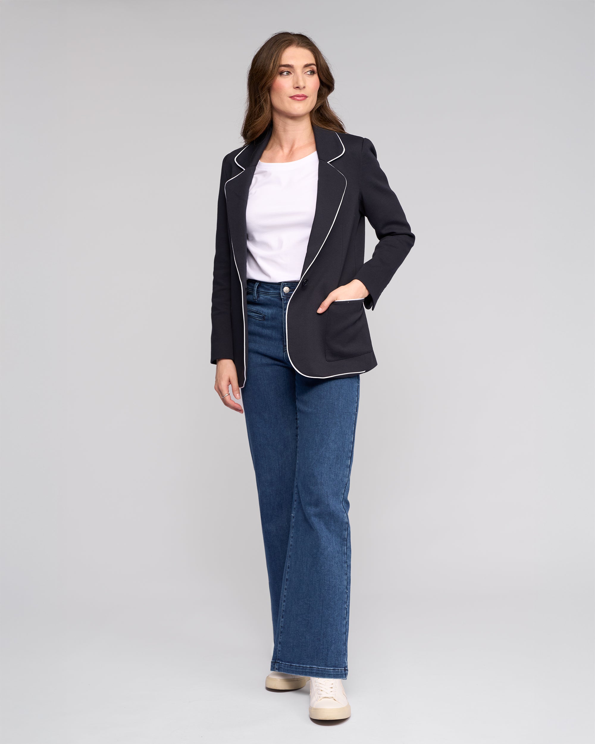 A woman with long brown hair poses confidently against a plain background, wearing the Vassalli Ora Blazer in Ink with white trim, a white shirt, high-waisted blue jeans, and white sneakers.