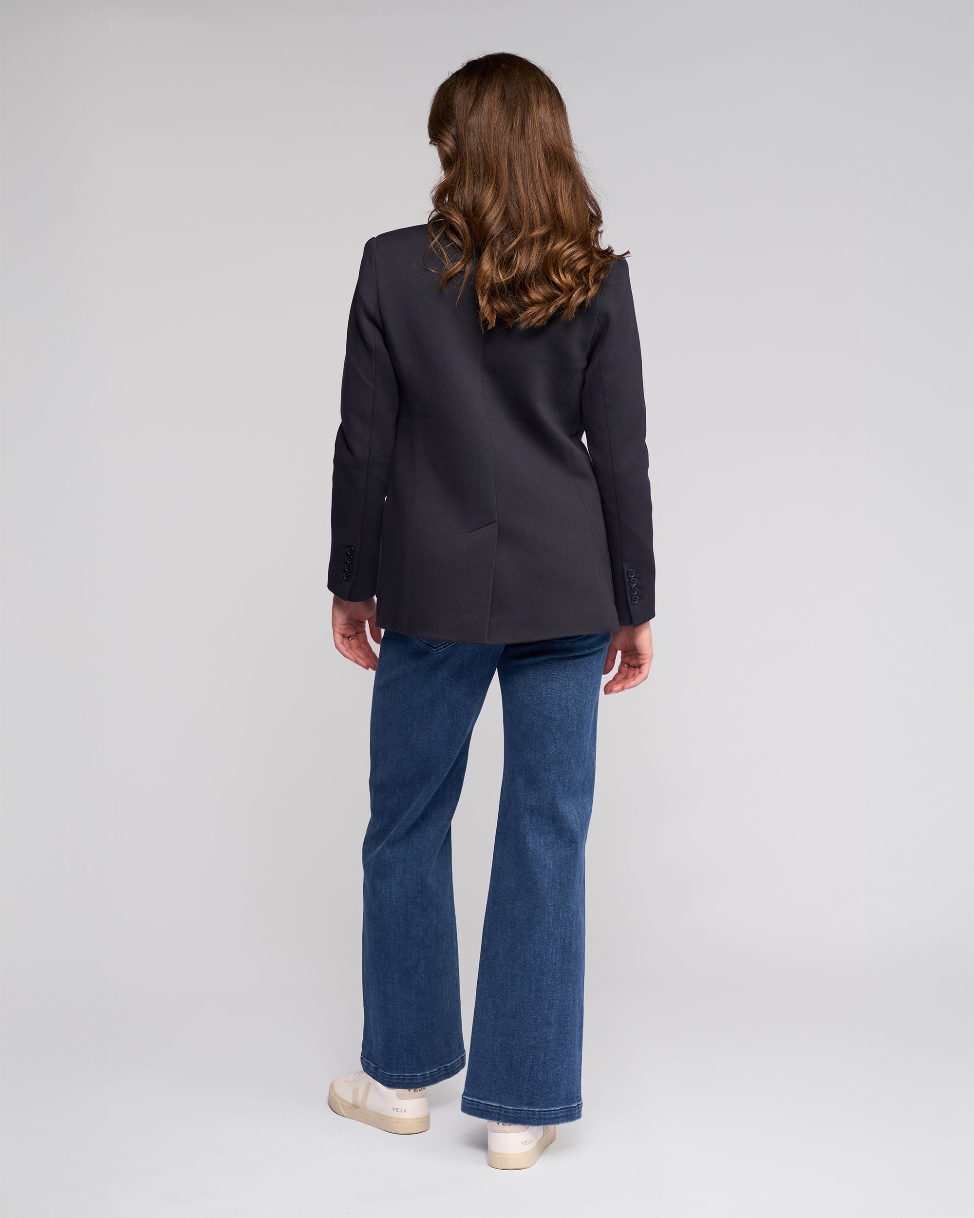 A person with long brown hair stands facing away, wearing the Vassalli Ora Blazer - Ink, blue jeans, and beige shoes against a plain light gray background.