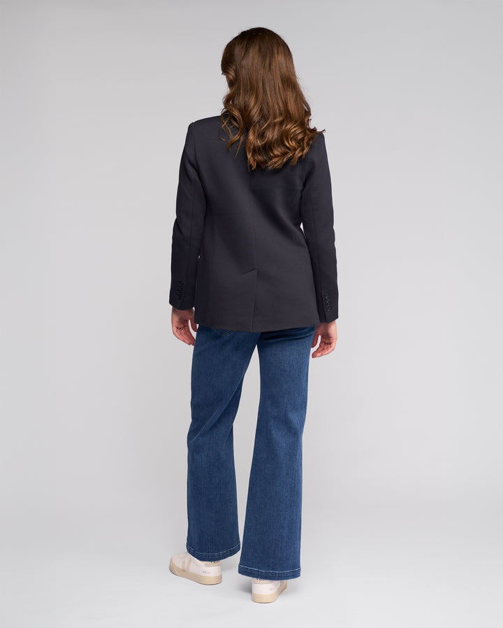 A person with long brown hair stands facing away, wearing the Vassalli Ora Blazer - Ink, blue jeans, and beige shoes against a plain light gray background.