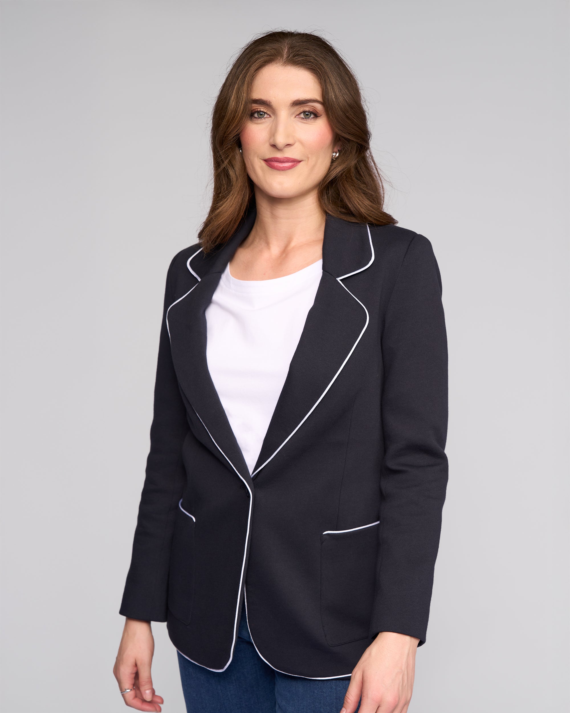 A woman with long brown hair smiles softly at the camera, wearing the Vassalli Ora Blazer in Ink over a cotton jersey top and blue jeans, set against a plain light gray background.