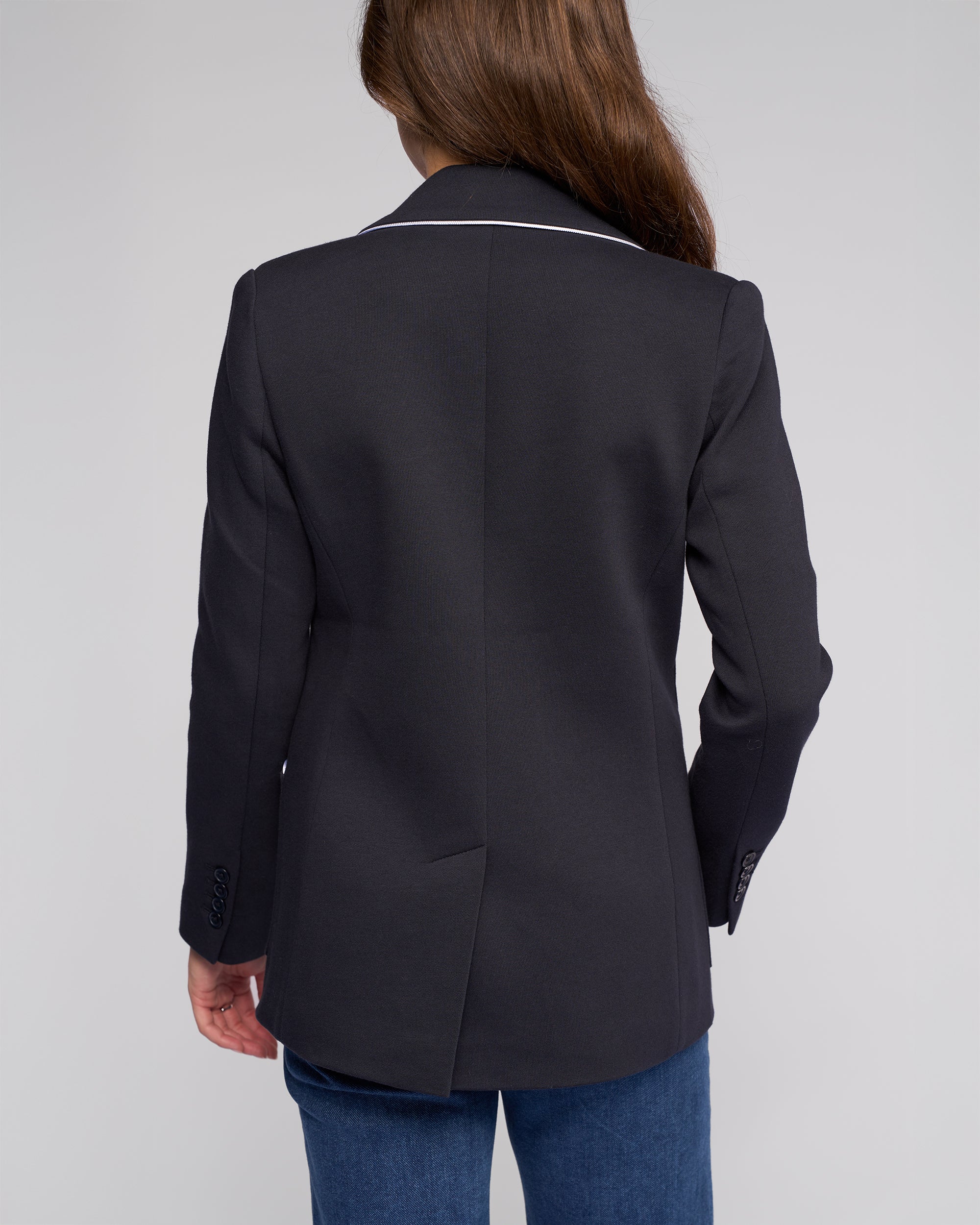 A person with long brown hair, shown from behind against a plain light background, is wearing the Vassalli Ora Blazer - Ink and blue jeans.