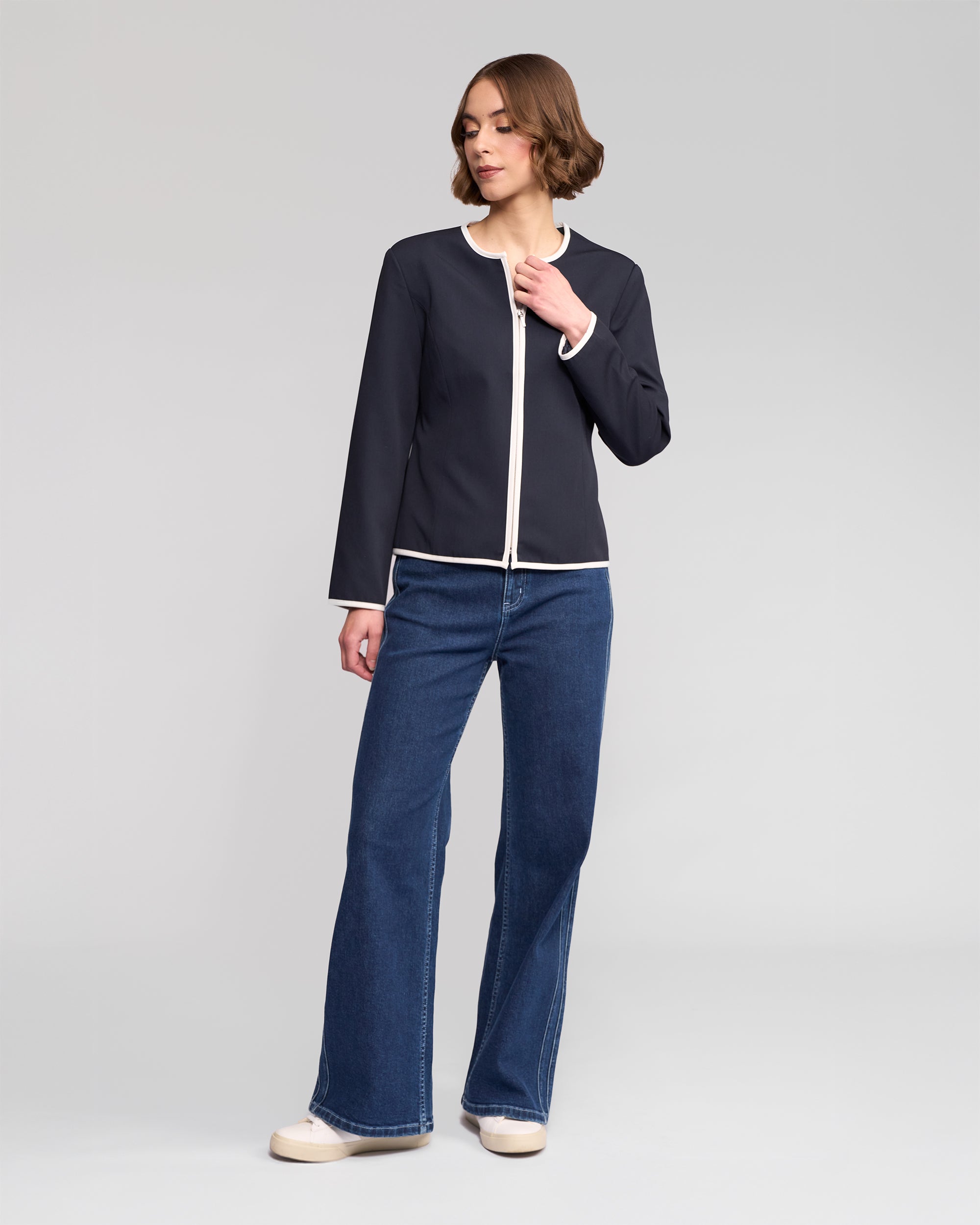 A person models the Vassalli Pier Jacket in Ink/White, featuring white trim, with blue flared jeans and white shoes, standing against a plain light gray background for a streamlined look.