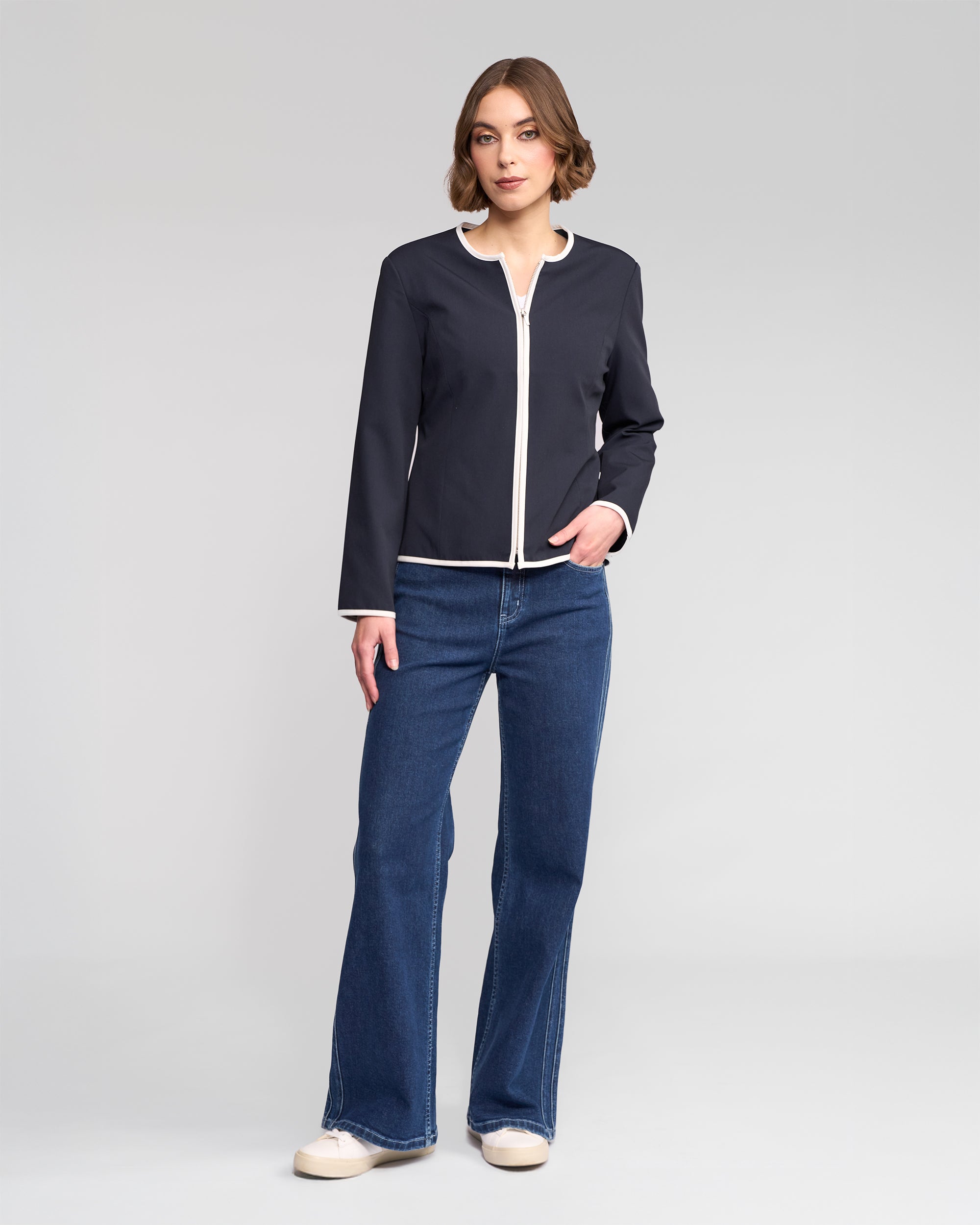 A woman stands in a neutral pose wearing the Vassalli Pier Jacket in Ink/White, paired with blue jeans and white sneakers, highlighting a streamlined silhouette against a plain light gray background.