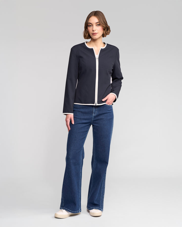 A woman stands in a neutral pose wearing the Vassalli Pier Jacket in Ink/White, paired with blue jeans and white sneakers, highlighting a streamlined silhouette against a plain light gray background.