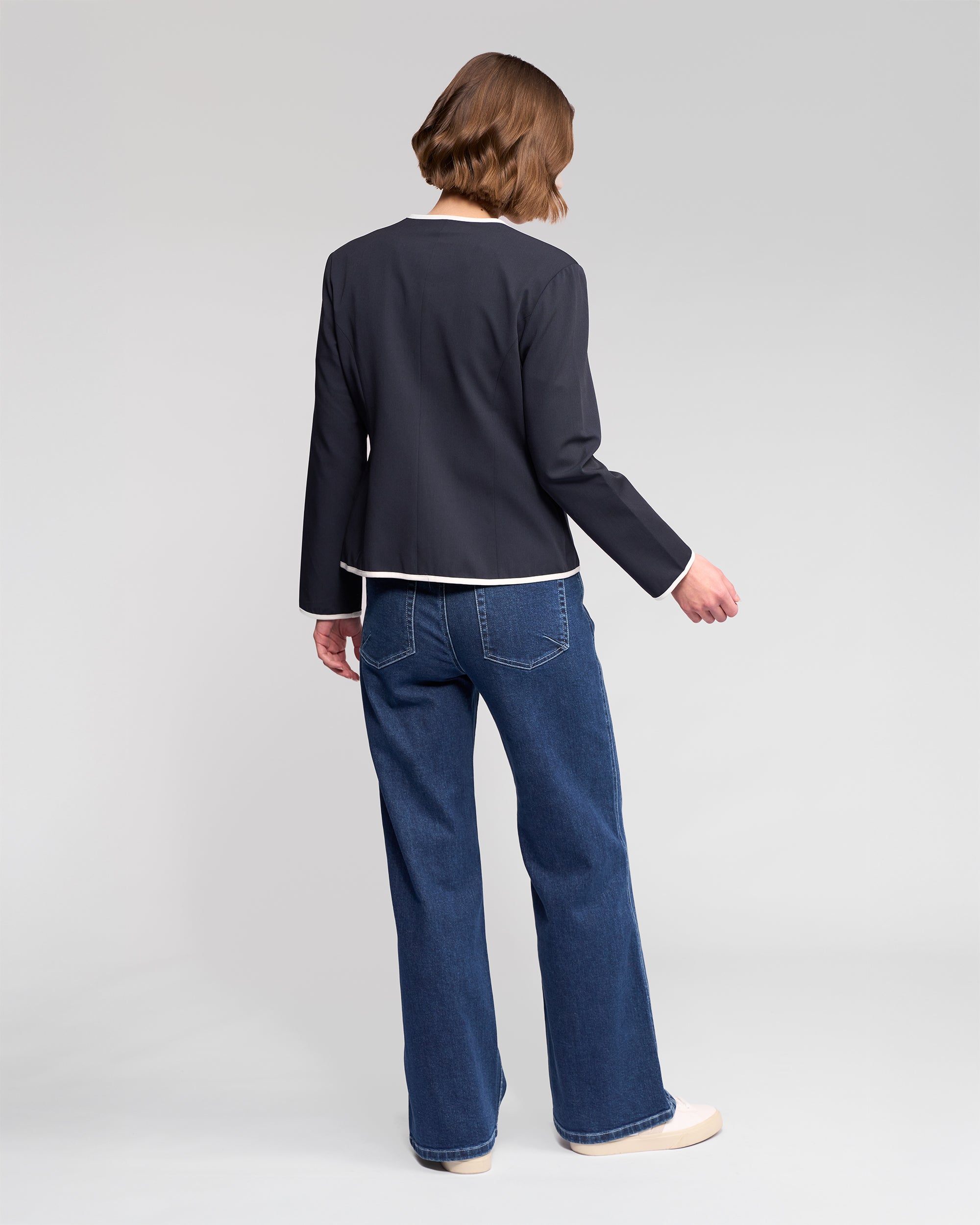 A person with short brown hair, wearing the Vassalli Pier Jacket - Ink/White, a white top, blue jeans, and beige shoes stands facing away from the camera against a plain light gray background.