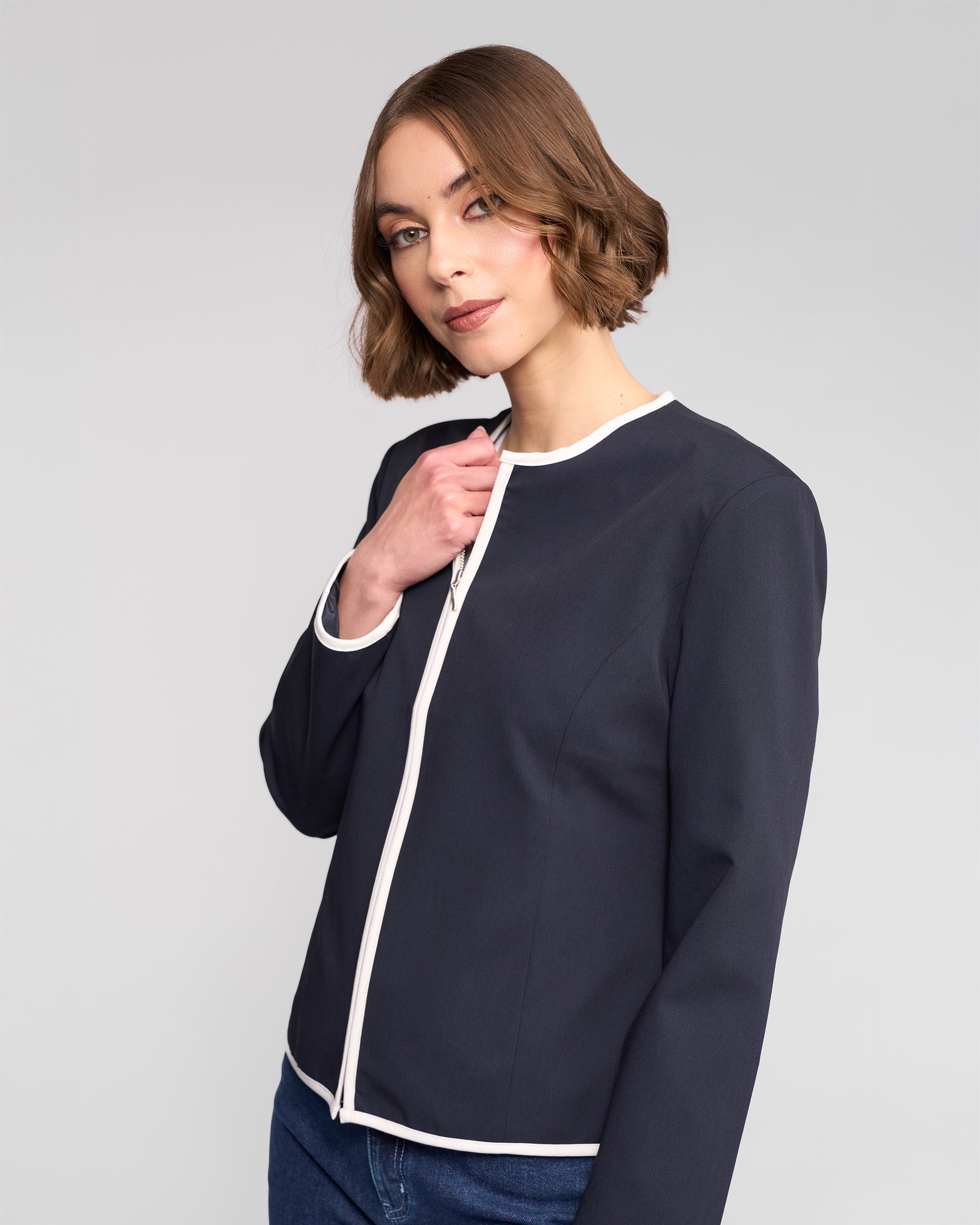 A woman with short brown hair wears the Vassalli Pier Jacket in Ink/White and blue jeans, standing confidently against a plain light gray background.