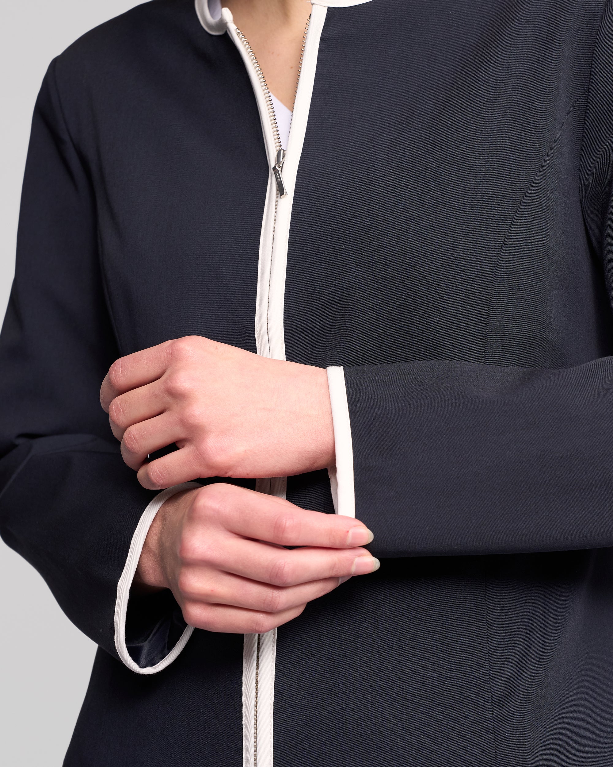 A person adjusts the cuff of their Vassalli Pier Jacket—Ink/White, a streamlined navy zip-up with crisp white trim and zipper. Their head is not visible.