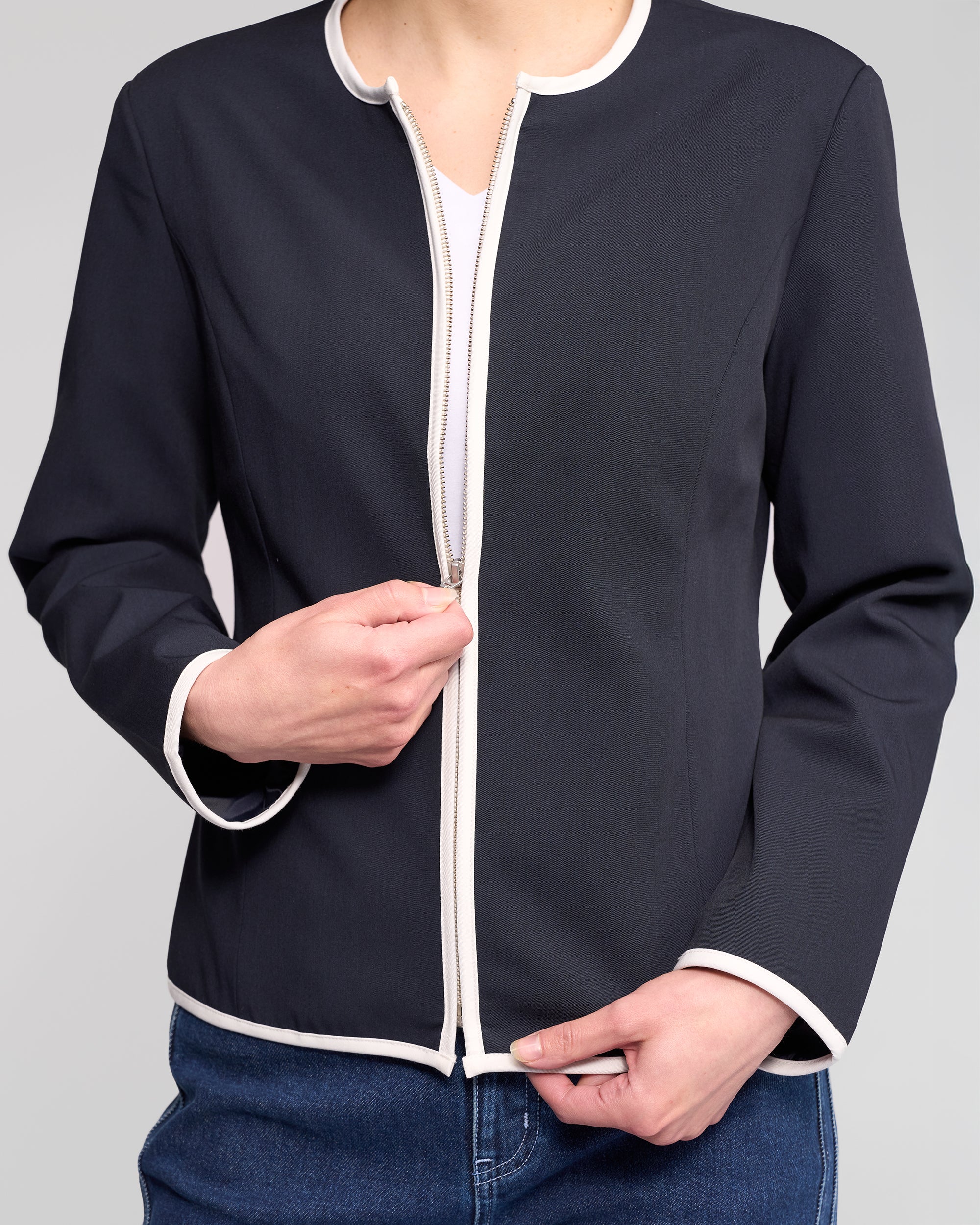 A person models the Vassalli Pier Jacket in Ink/White, partially unzipped to reveal a white shirt, paired with blue jeans. The streamlined silhouette is highlighted while the model’s face remains out of frame.