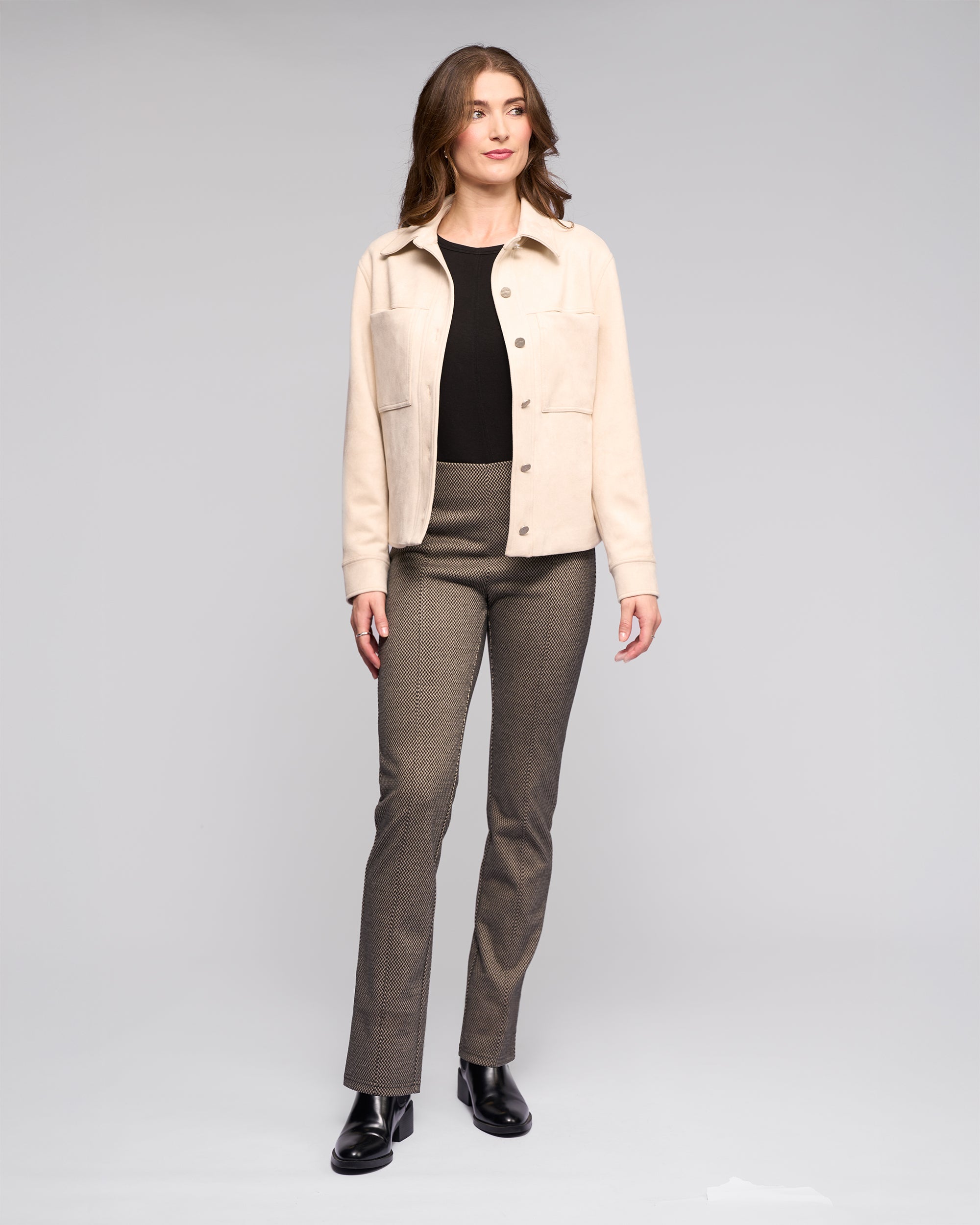 A woman stands facing forward in the Vassalli Guest Jacket - Bone, styled with a black top, grayish-brown pants, and black shoes against a plain light gray background.