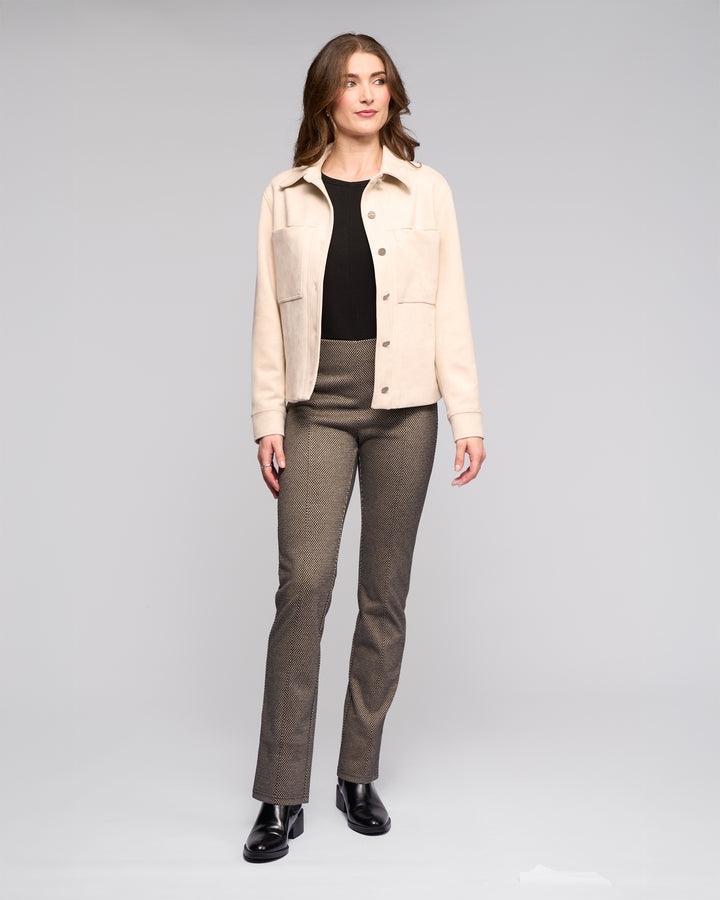 A woman stands facing forward in the Vassalli Guest Jacket - Bone, styled with a black top, grayish-brown pants, and black shoes against a plain light gray background.