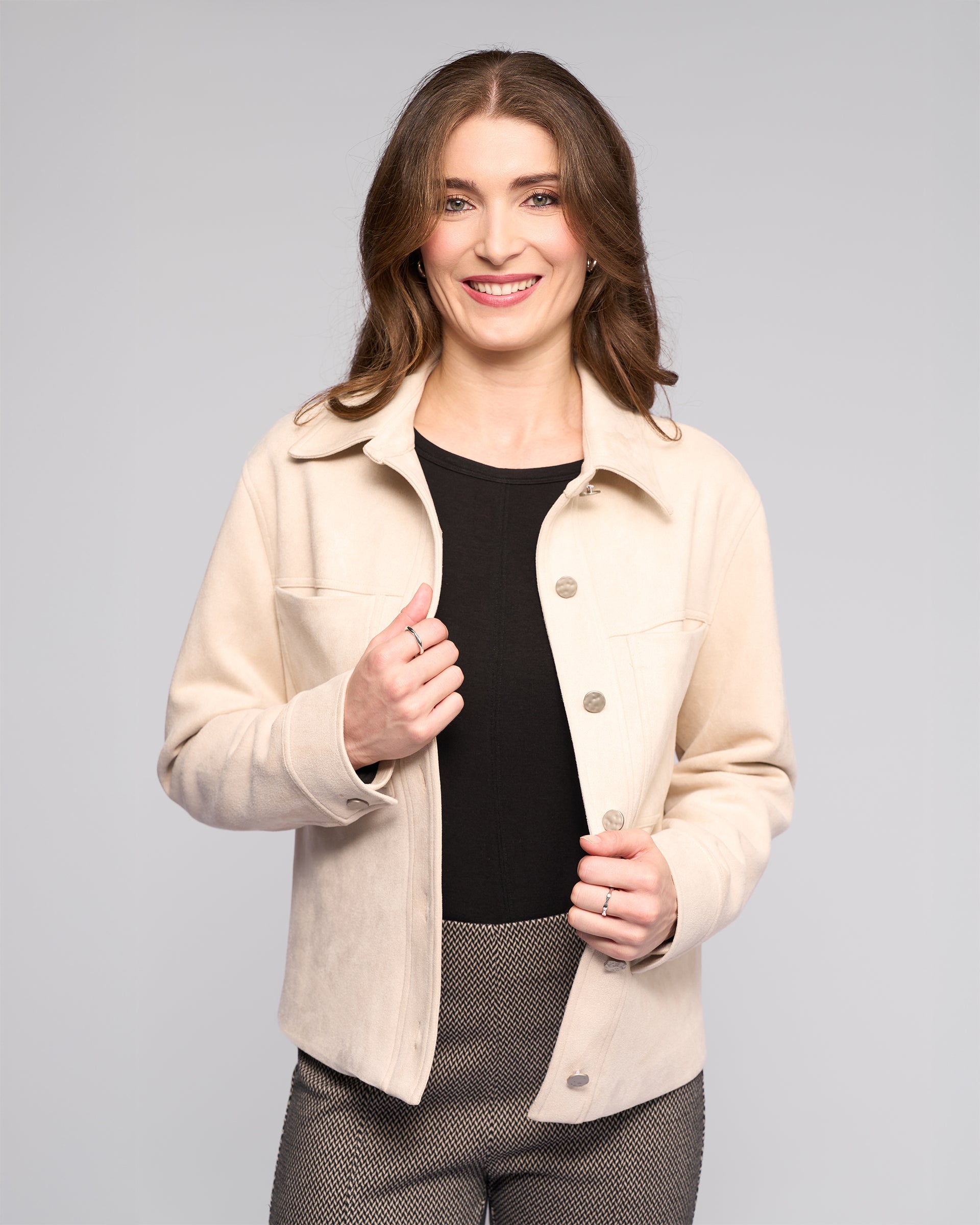 A woman with long brown hair smiles at the camera, wearing the Vassalli Guest Jacket in Bone over a black top and patterned pants, standing against a light gray background.