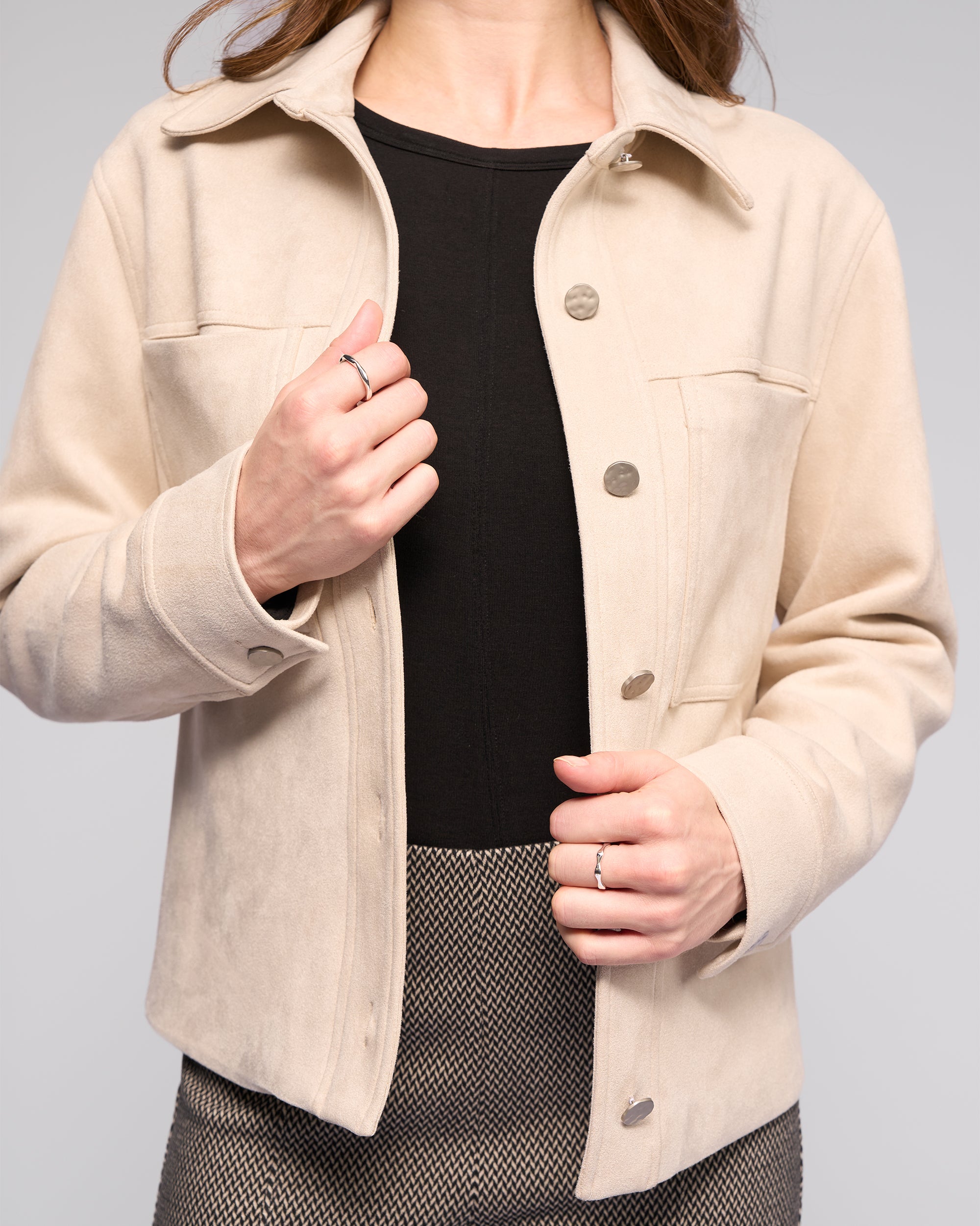 A woman wears the Vassalli Guest Jacket - Bone over a black top and herringbone skirt, adjusting the jacket with one hand while the other rests near the buttons. Only her torso and hands are visible.