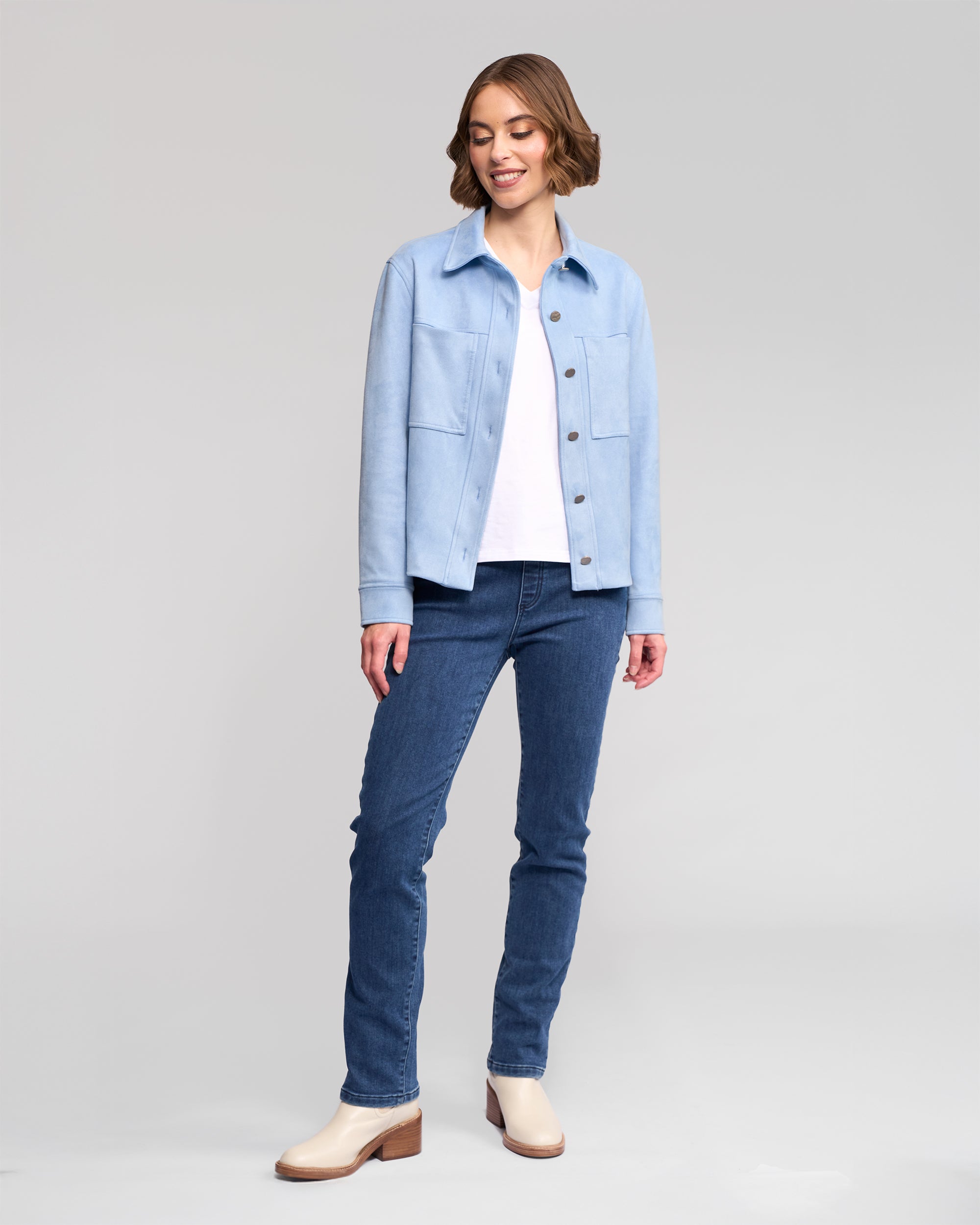A woman smiles, wearing the Vassalli Guest Jacket in sky blue over a white shirt, blue jeans, and light-colored boots against a plain light gray background.