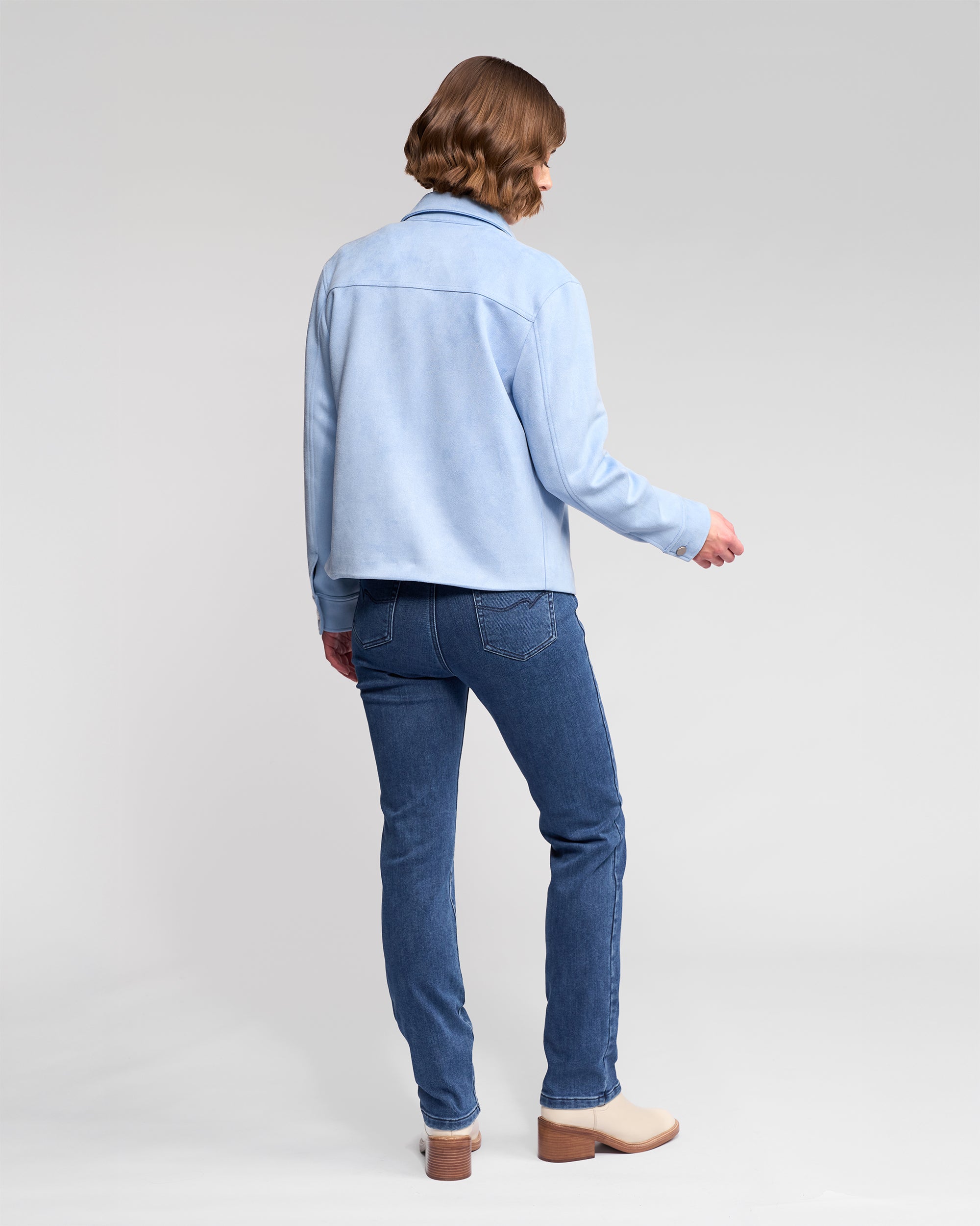 Wearing the Vassalli Guest Jacket in Sky Blue, a person with short, wavy brown hair and blue jeans stands facing away from the camera against a plain light backdrop.