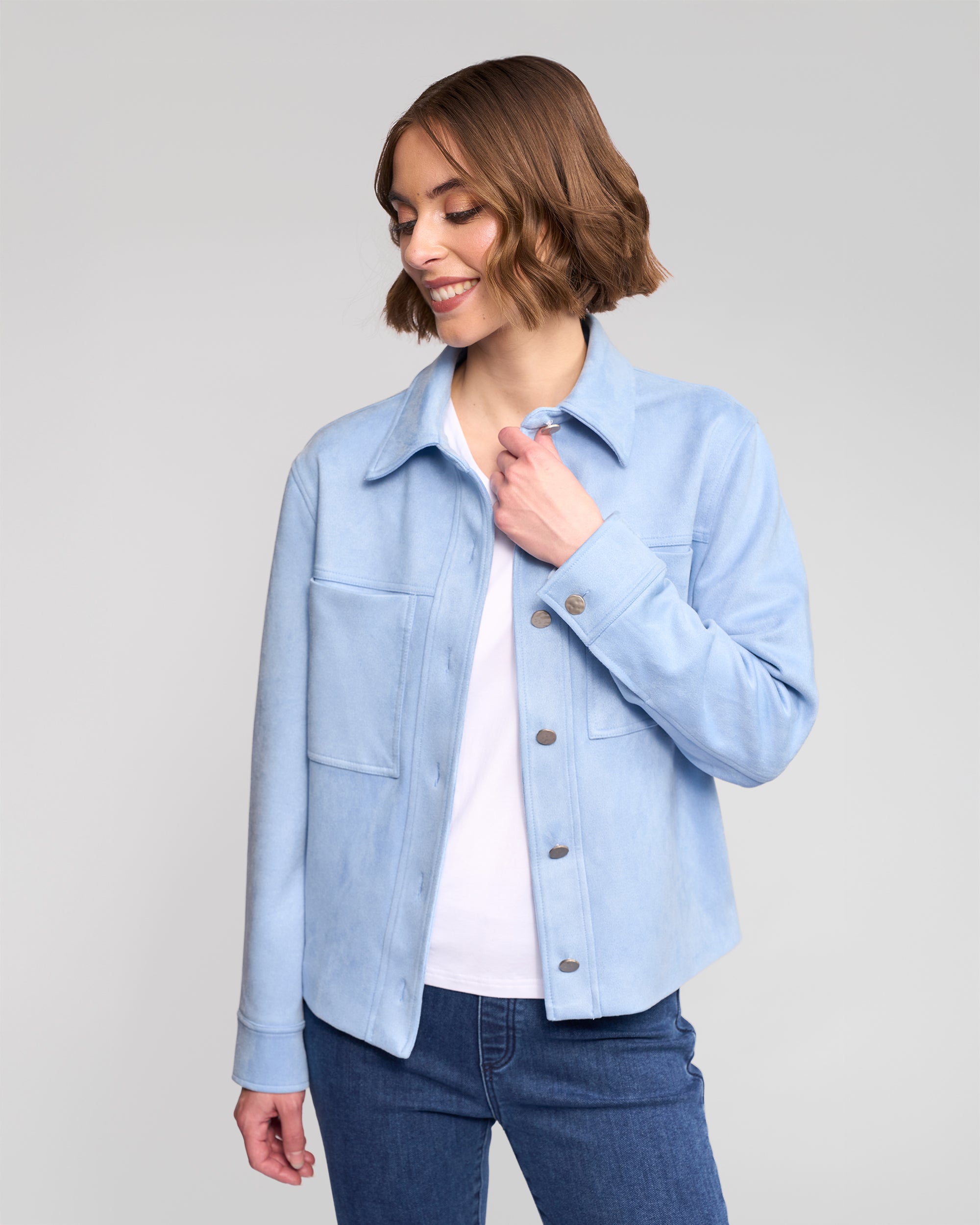 A woman with short brown hair smiles while wearing the Vassalli Guest Jacket in Sky Blue over a white shirt and blue jeans, standing against a light gray background.