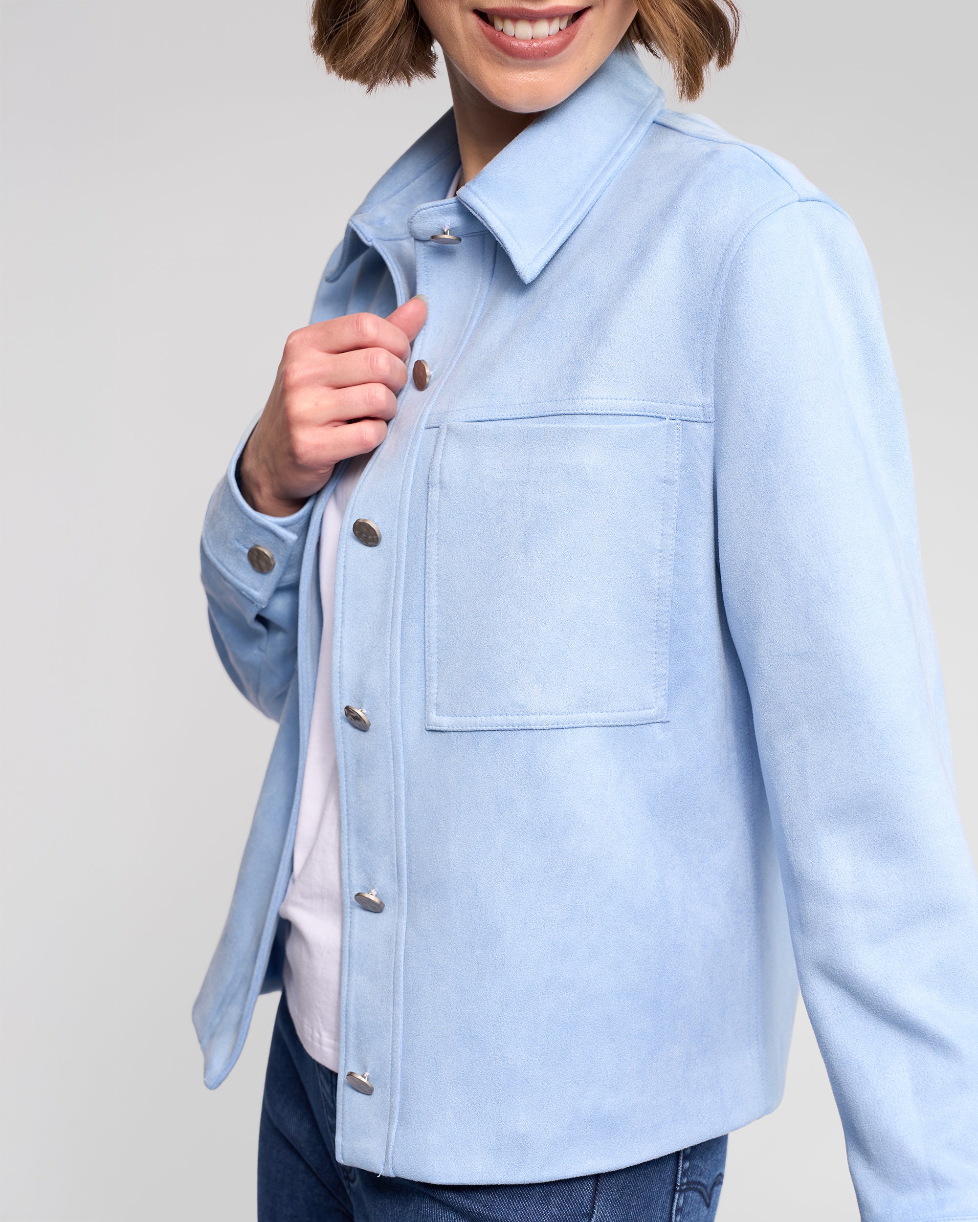 A woman in the Vassalli Guest Jacket - Sky Blue over a white shirt and blue jeans smiles, holding the jacket’s collar. Her face is partially out of frame.