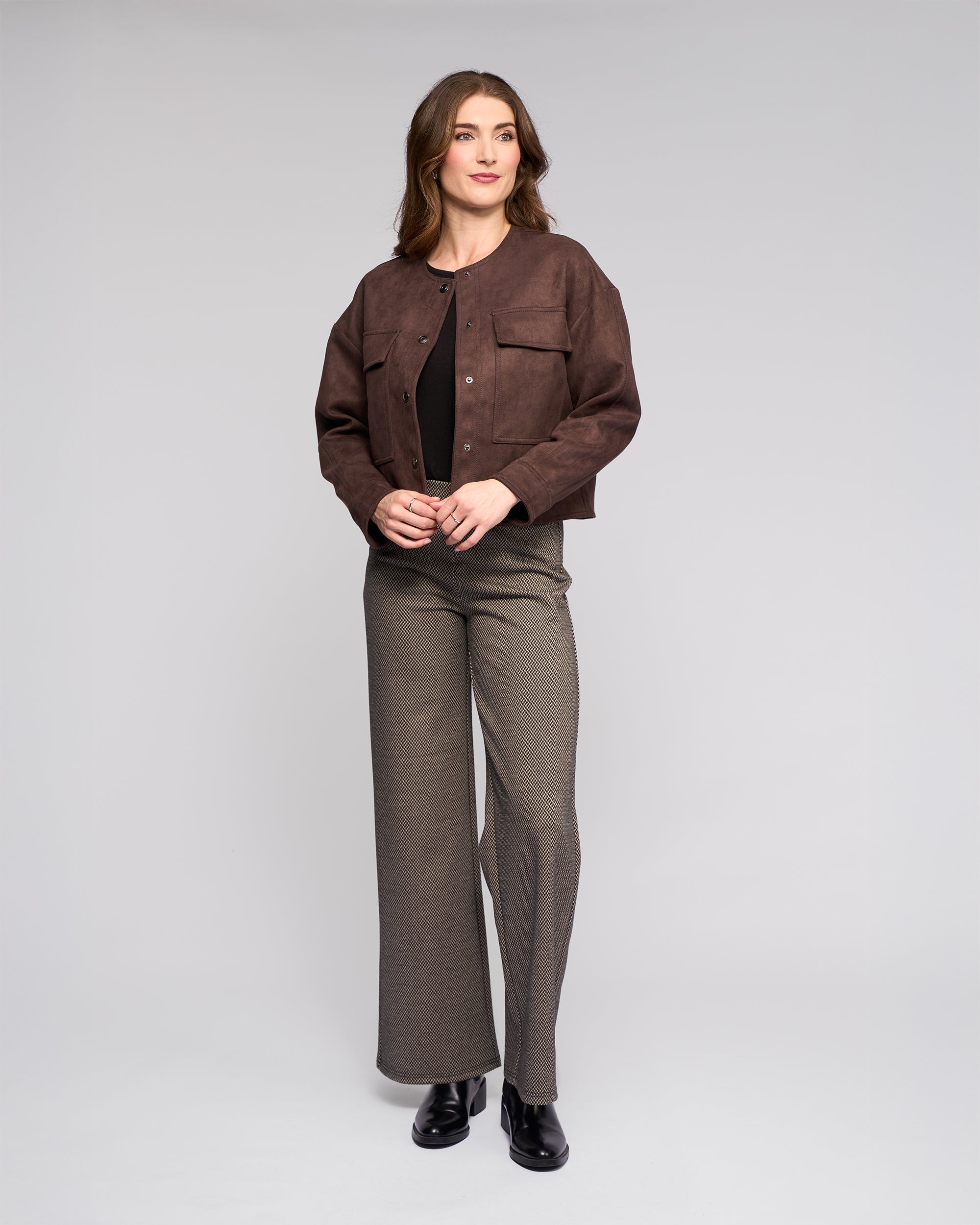 A woman with wavy brown hair and a neutral expression stands against a light gray background, wearing the Vassalli Relay Jacket in chocolate, a dark top, gray wide-leg pants, and black shoes, with her hands lightly clasped.