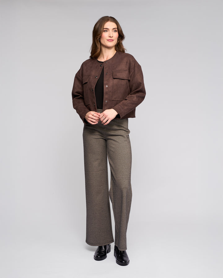 A woman with wavy brown hair and a neutral expression stands against a light gray background, wearing the Vassalli Relay Jacket in chocolate, a dark top, gray wide-leg pants, and black shoes, with her hands lightly clasped.