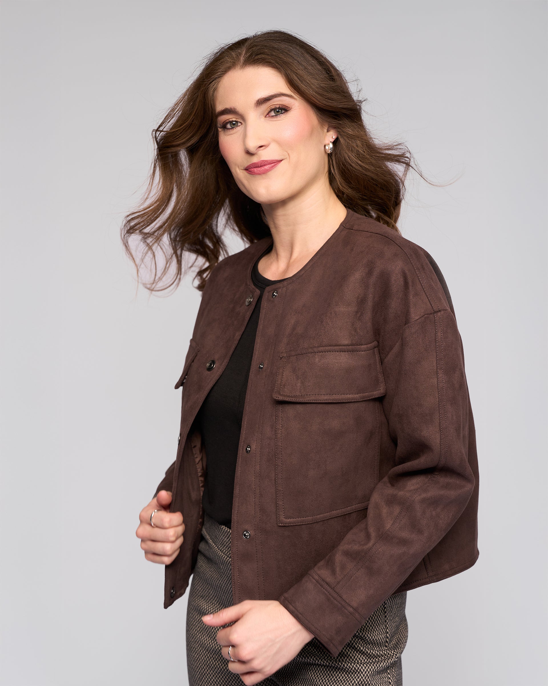 A woman with long brown hair models the Vassalli Relay Jacket in chocolate, styled over a black top and patterned pants, smiling confidently against a light gray background.