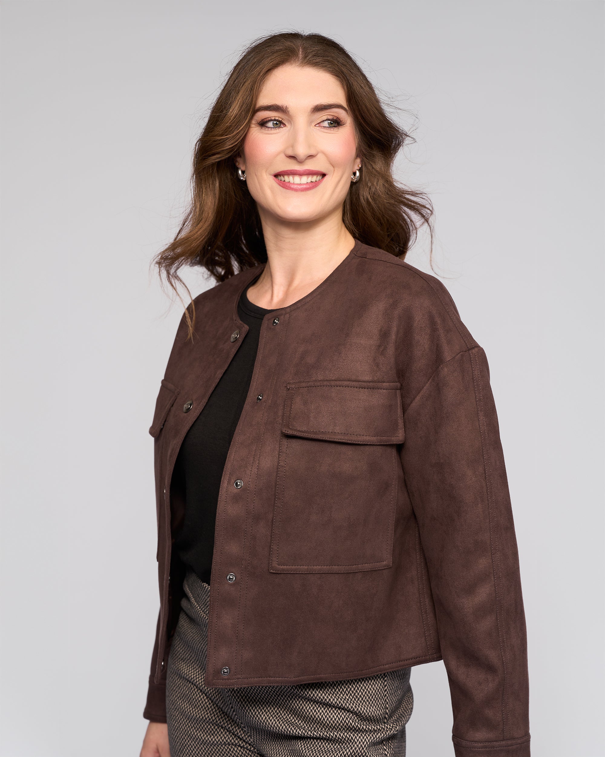 A woman with long brown hair smiles and looks to the side, wearing the Vassalli Relay Jacket in Chocolate over a black top and patterned pants against a plain light gray background.