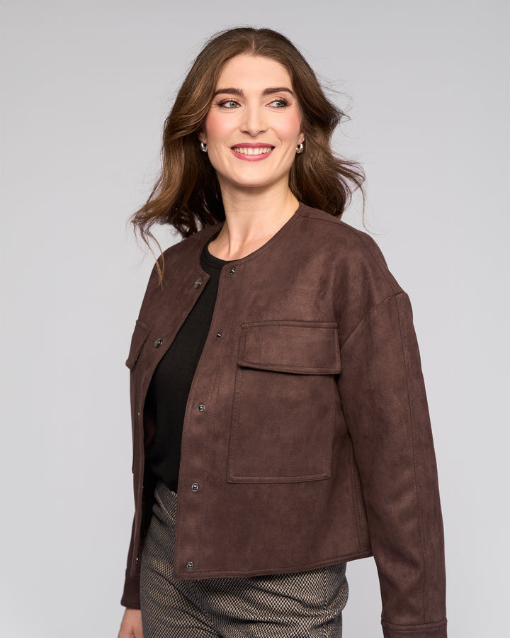 A woman with long brown hair smiles and looks to the side, wearing the Vassalli Relay Jacket in Chocolate over a black top and patterned pants against a plain light gray background.