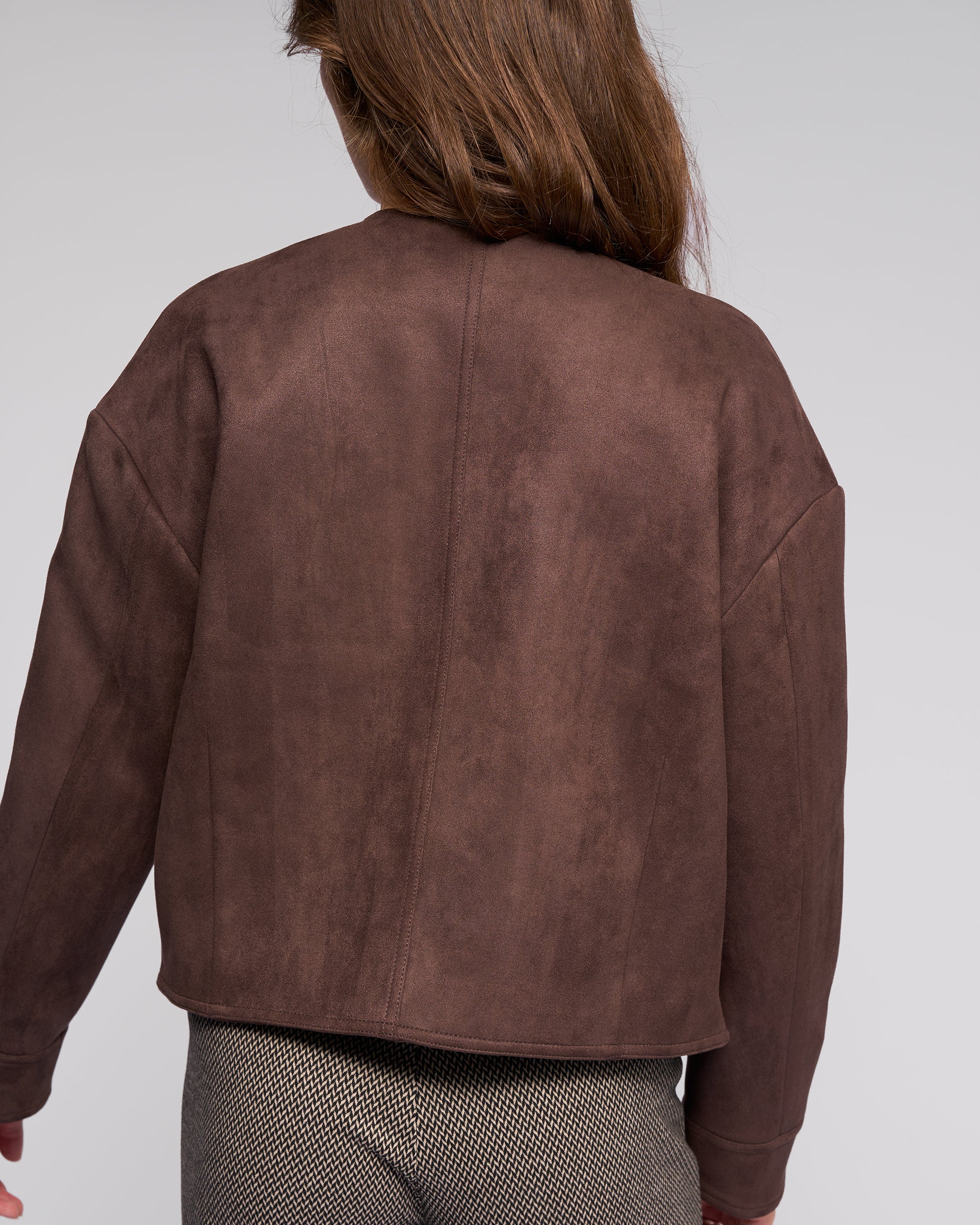 A person with long brown hair is seen from the back wearing the Vassalli Relay Jacket in chocolate and textured patterned pants, set against a plain light background.
