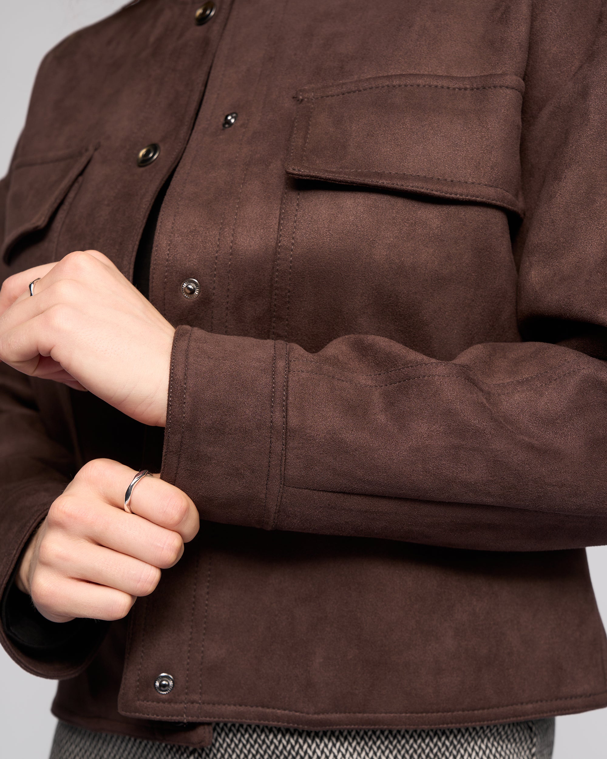 A person fastens the cuff of the Vassalli Relay Jacket - Chocolate, featuring snap buttons. Only the hands and upper torso are visible, with a silver ring on one finger.