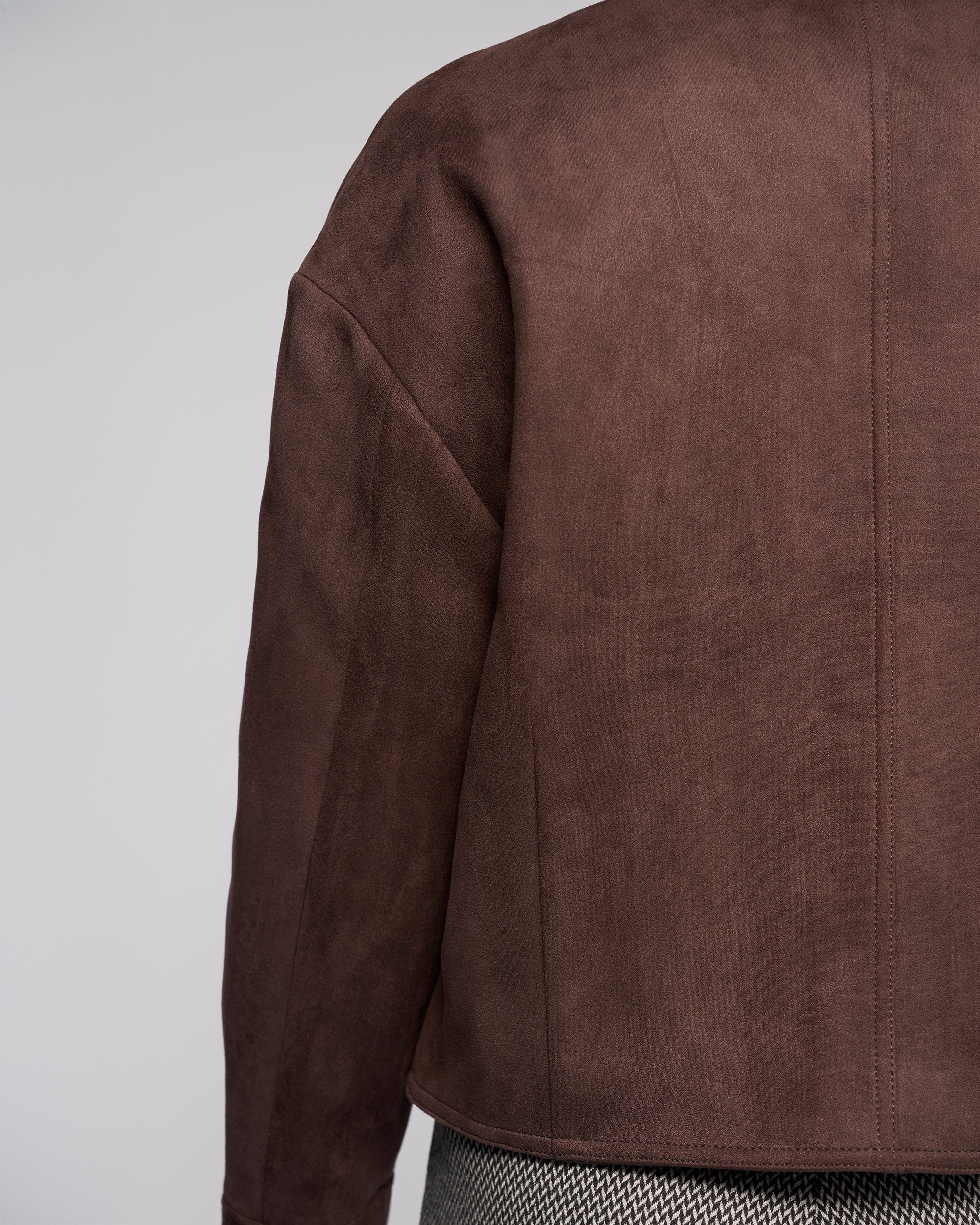 A close-up of the back of a person wearing the Vassalli Relay Jacket in Chocolate and herringbone-patterned pants, set against a plain, light-colored background.