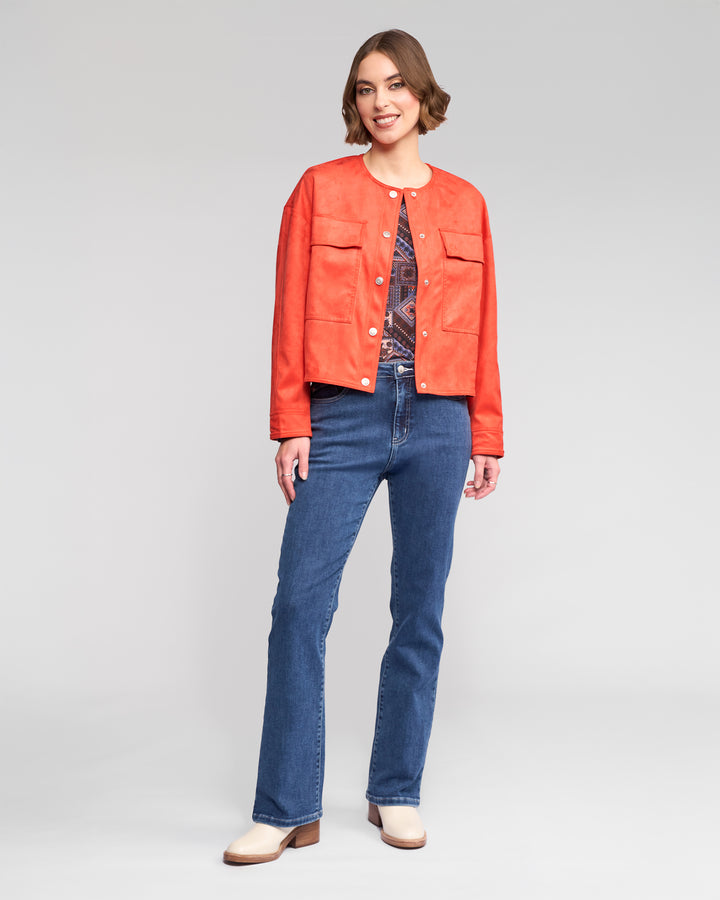 A woman smiles in the Vassalli Relay Jacket - Lava, a bold orange collarless faux suede jacket, worn over a patterned top with blue jeans and cream shoes against a plain light grey background.