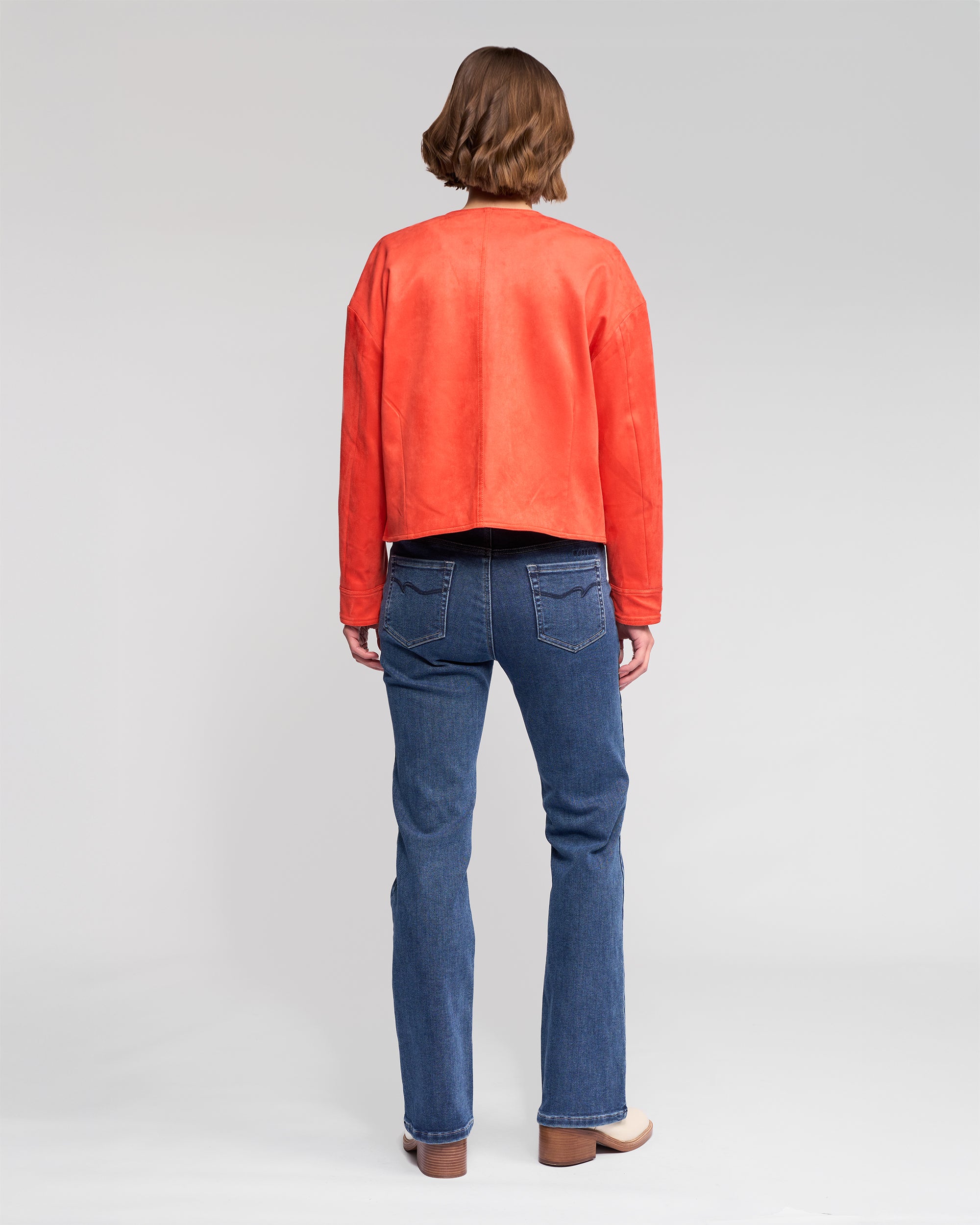 A person with short brown hair, wearing the Vassalli Relay Jacket - Lava, blue jeans, and brown shoes stands facing away against a plain light gray background.