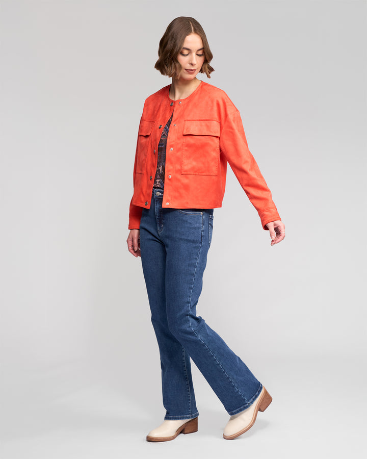 A woman with short brown hair models the Vassalli Relay Jacket in Lava, pairing it with blue jeans and beige heeled shoes. She stands against a plain light gray background, looking down with one arm slightly extended.