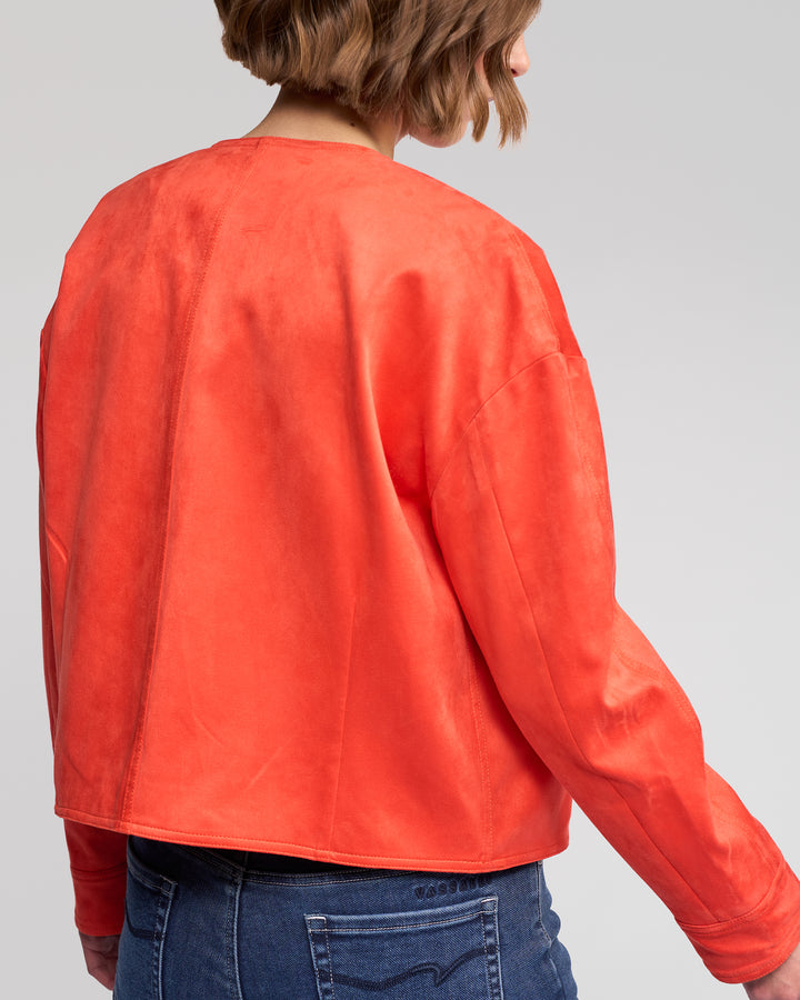 A person with short, wavy brown hair wears the Vassalli Relay Jacket - Lava, a bold orange faux suede design without a collar, paired with blue denim jeans and shown from the back against a plain light gray background.