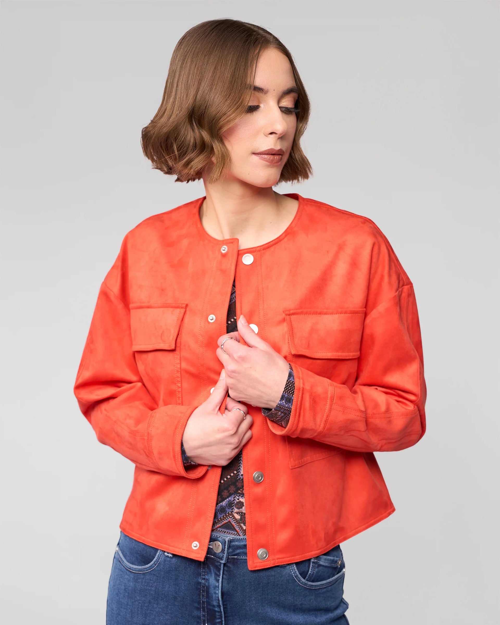 A woman with short brown hair wears the Vassalli Relay Jacket in Lava over a patterned top and blue jeans. She stands against a plain light gray background, looking down and slightly to the side.