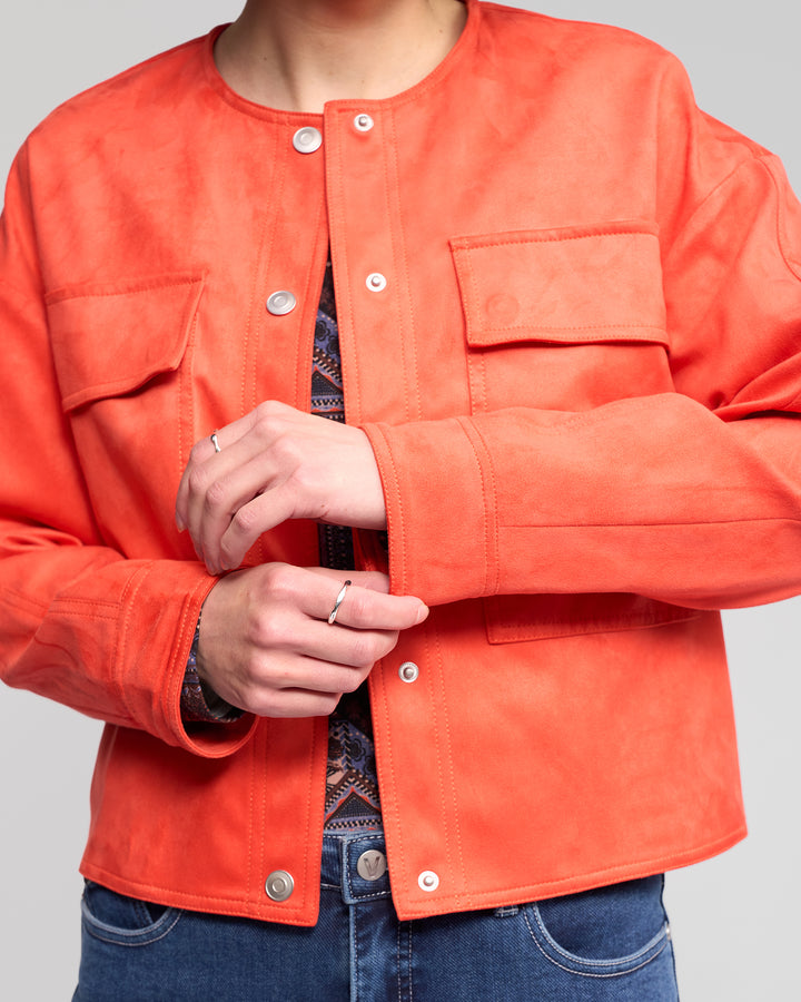 A person wearing the Vassalli Relay Jacket - Lava, a bold orange collarless faux suede jacket with snap buttons and chest pockets, adjusts their sleeve. The jacket is styled over a patterned top, blue jeans, and rings.