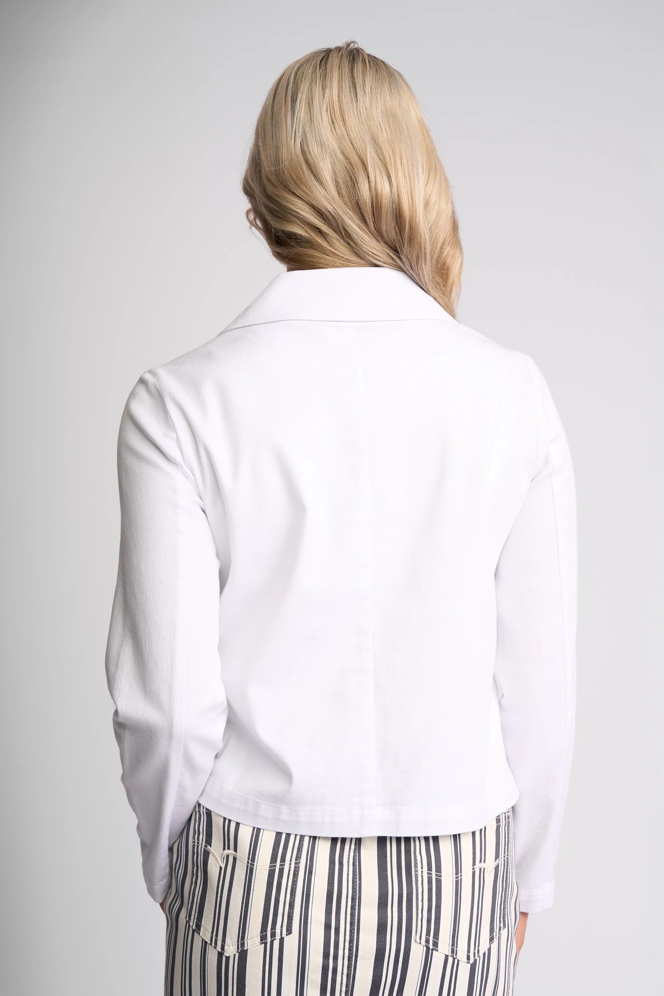 A person with long blonde hair, seen from behind, wears the Vassalli Hera Jacket in white denim and a white skirt with black vertical stripes, standing against a plain light background.