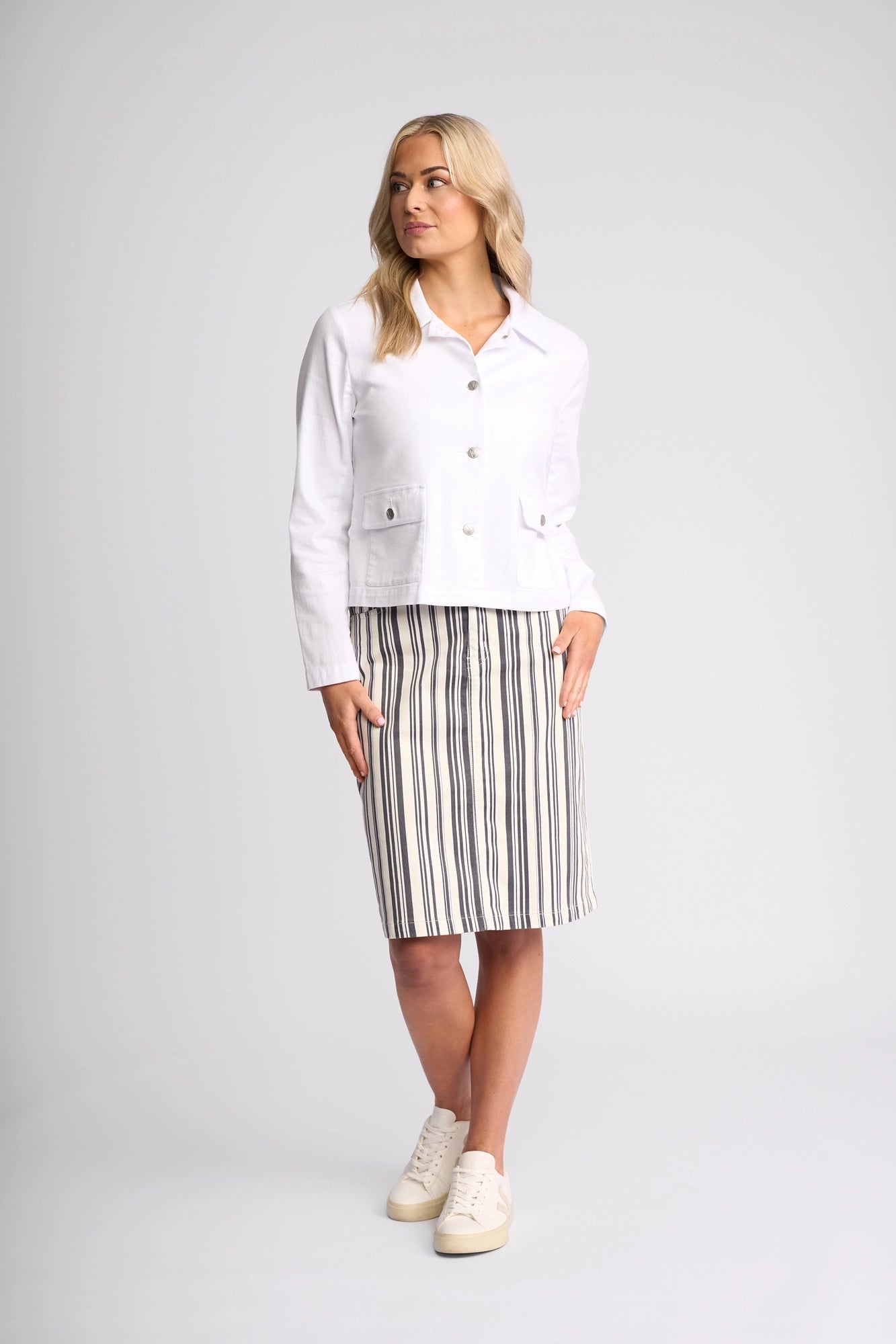 A woman wearing the Vassalli Hera Jacket in white denim, paired with a knee-length skirt featuring vertical black and white stripes and white sneakers, stands against a plain, light background.