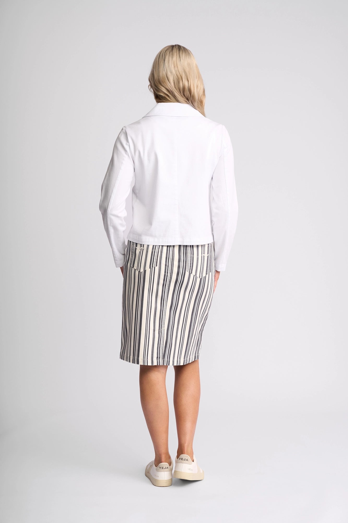 A person with blonde hair stands facing away, wearing the Vassalli Hera Jacket in white denim layered over a knee-length skirt featuring black and white vertical stripes, paired with white slip-on shoes, against a plain white background.