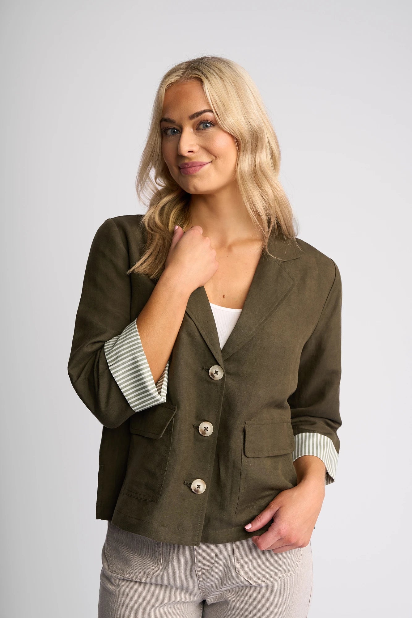 A woman with long blonde hair wears the Vassalli Honey Jacket in Khaki—a stylish olive green linen-viscose blazer with rolled sleeves—paired with a white top and light gray pants, standing against a plain light background and smiling softly.