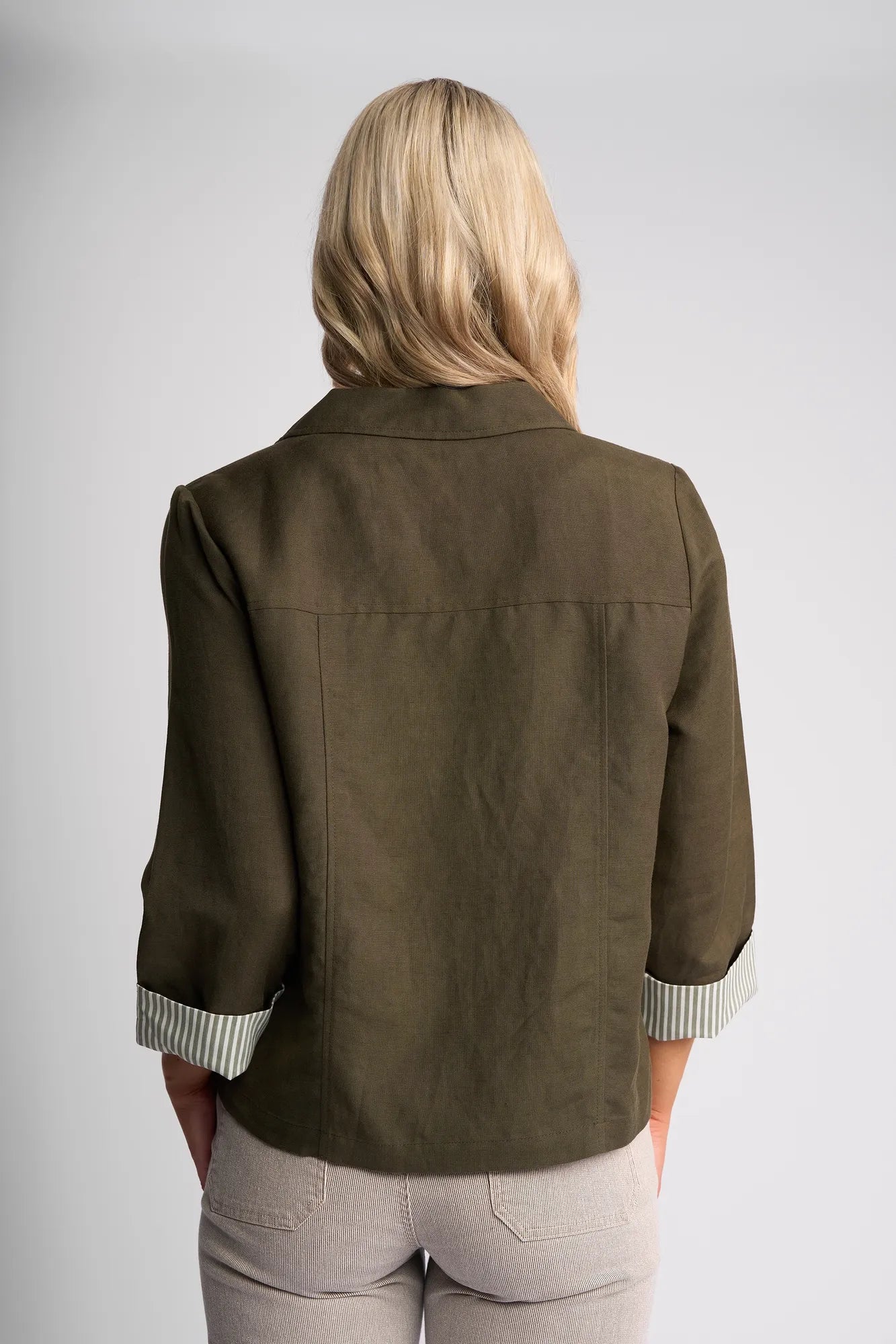 A woman with blonde hair is shown from the back, wearing a khaki Vassalli Honey Jacket with cuffed sleeves and beige pants, standing against a plain light gray background.