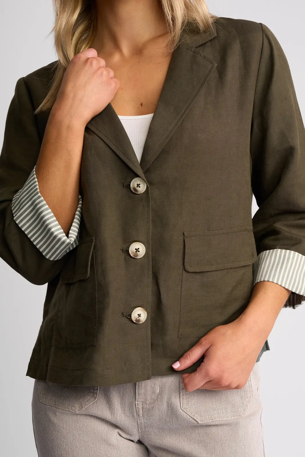 A woman wearing the Vassalli Honey Jacket in khaki—a lightweight linen-viscose piece with rolled sleeves revealing a striped lining—paired with a white top and light-colored pants, stands with one hand near her collar and the other in her pocket.
