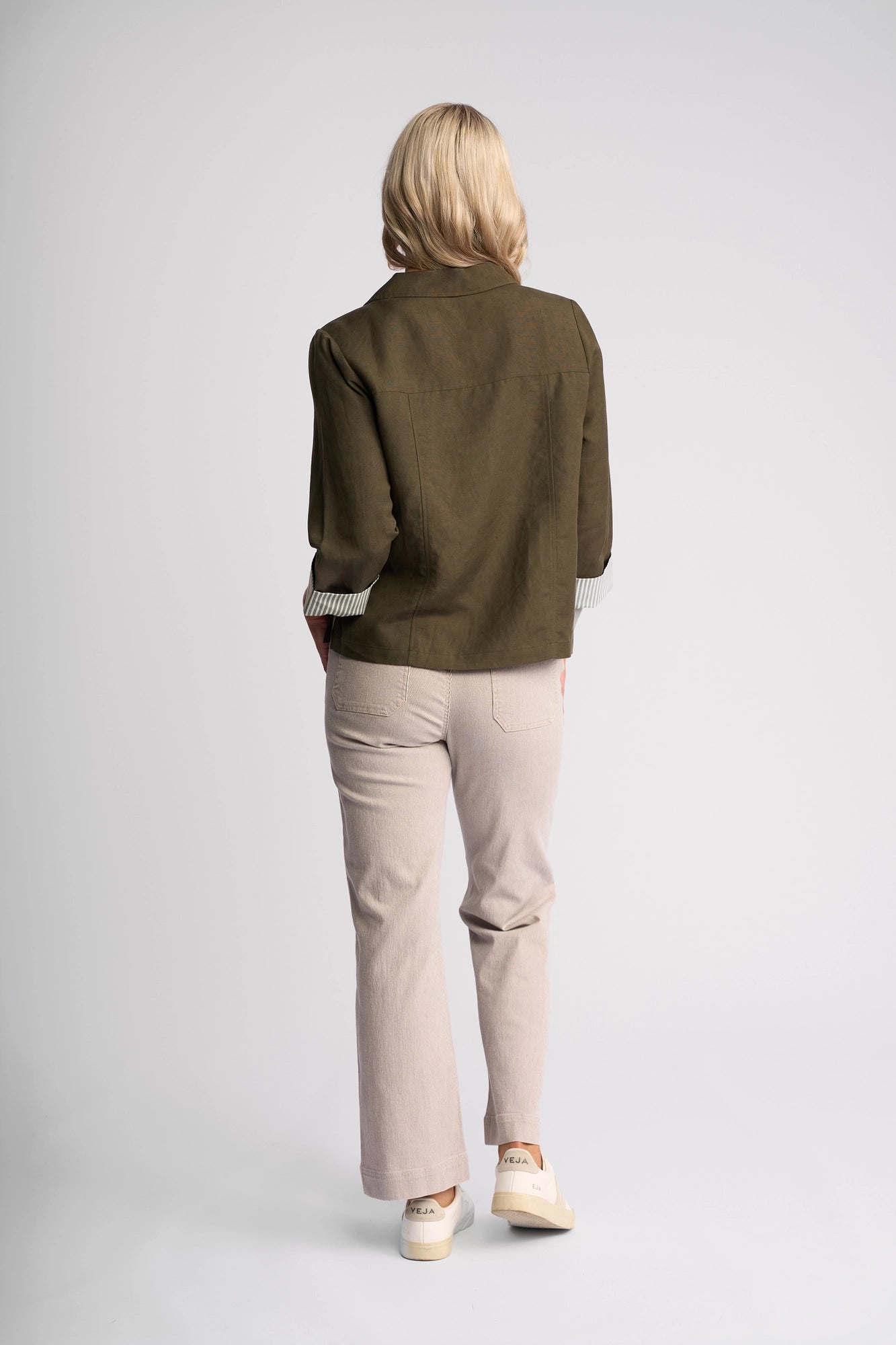 A person with shoulder-length blonde hair, dressed in the Vassalli Honey Jacket in Khaki, light-colored pants, and white sneakers, stands facing away from the camera against a plain white background.