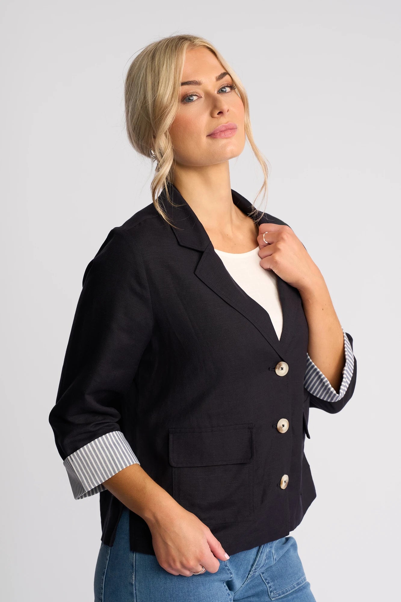 A woman with blonde hair in a low bun wears the Vassalli Honey Jacket in Navy, featuring lightweight linen-viscose fabric and rolled sleeves, styled with a white shirt and blue jeans, standing against a plain light background.