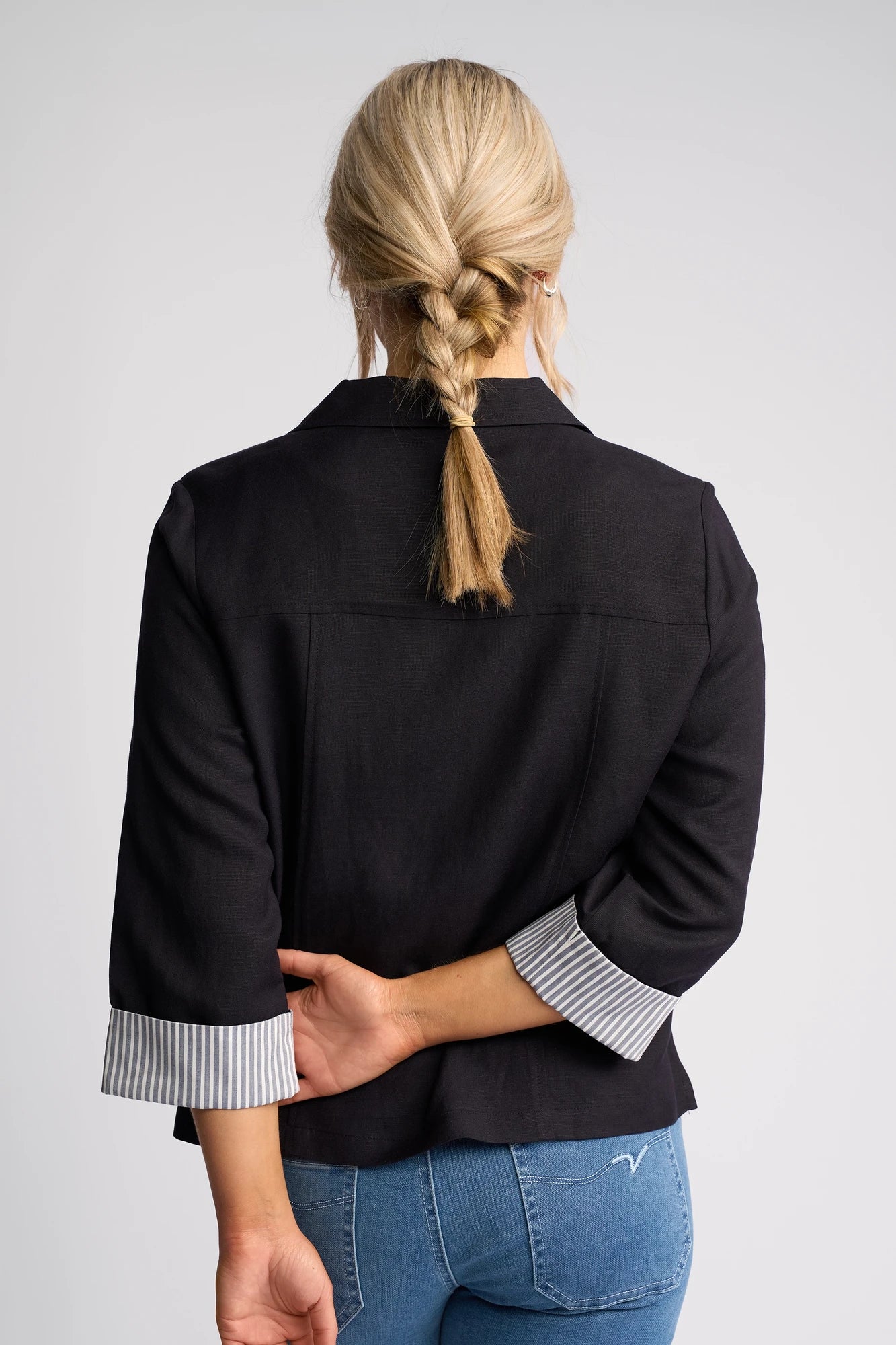 A woman with blonde hair in a braid stands with her back to the camera and one arm bent behind her back, wearing the Vassalli Honey Jacket in Navy with striped cuffs and blue jeans.