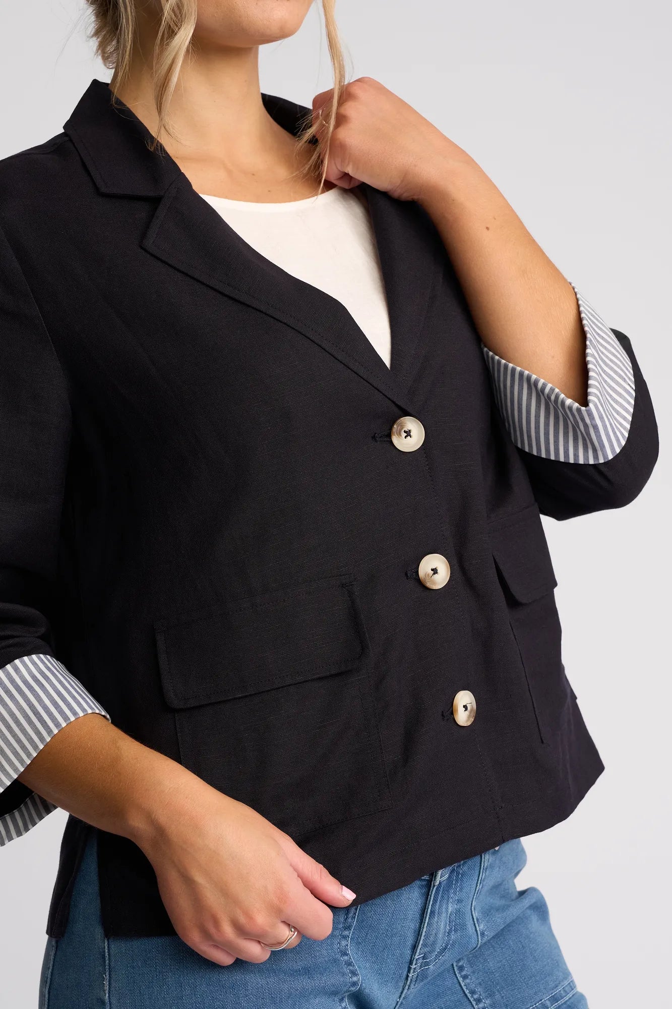 A woman wears the Vassalli Honey Jacket in navy, styled over a white top and blue jeans. With light hair partially pulled back, she poses with one hand near her collar and the other relaxed by her side, creating an effortlessly stylish look. The jacket’s striped sleeve lining subtly elevates the ensemble.
