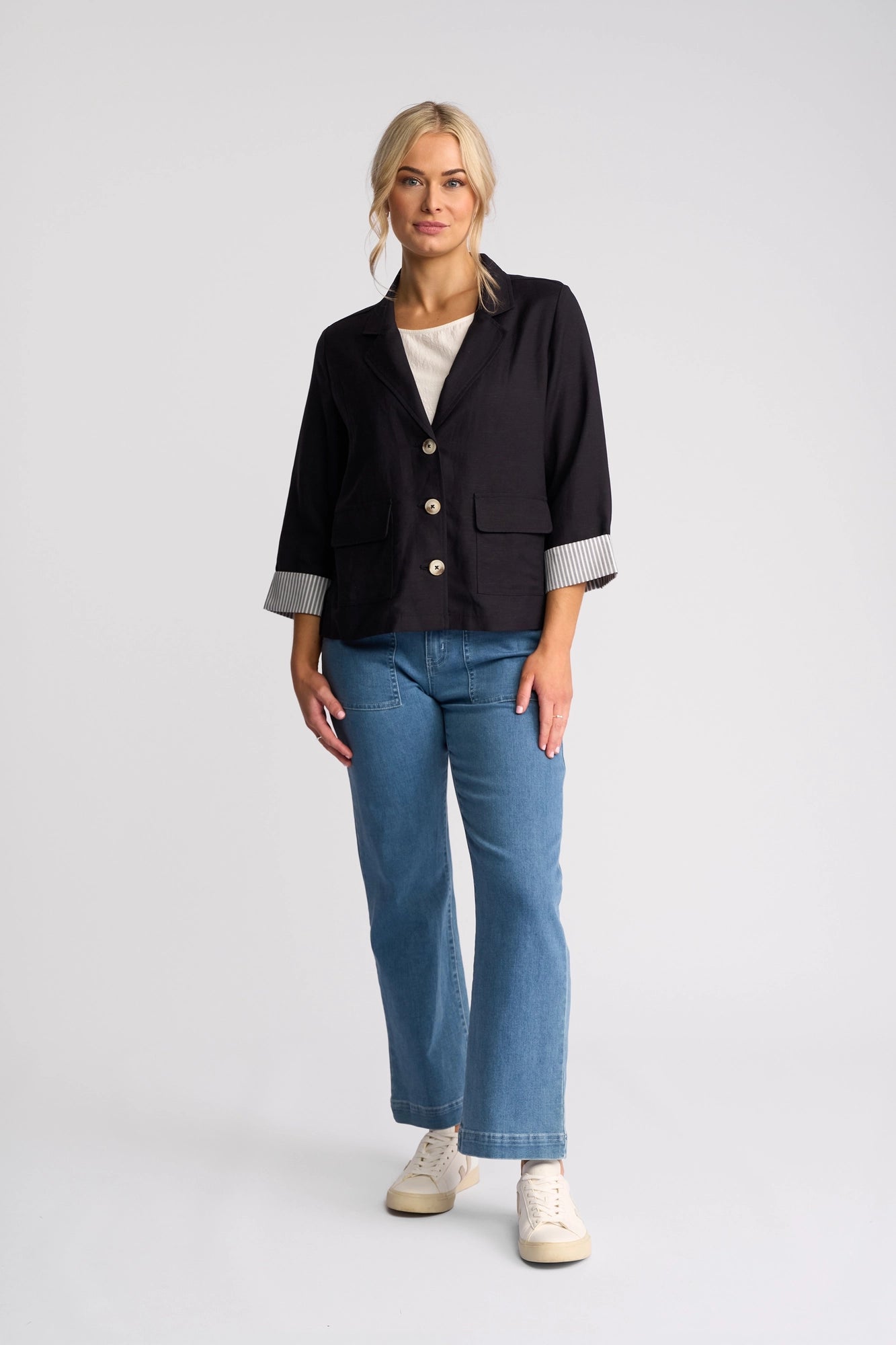 A woman stands facing forward, wearing the Vassalli Honey Jacket in navy, paired with a white top, blue jeans, and white sneakers, against a plain white background.