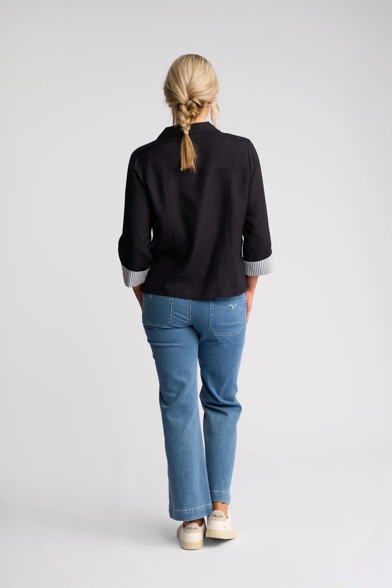 A person with blonde hair in a braid, wearing the Vassalli Honey Jacket in Navy, blue jeans, and white shoes stands facing away with hands in pockets against a plain white background.