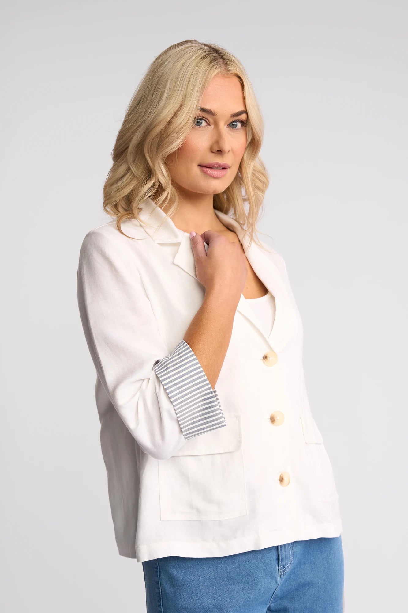 A woman with blonde hair wears the Vassalli Honey Jacket in white, a lightweight linen-viscose blazer featuring striped cuffs, paired with blue jeans as she stands against a plain light background.