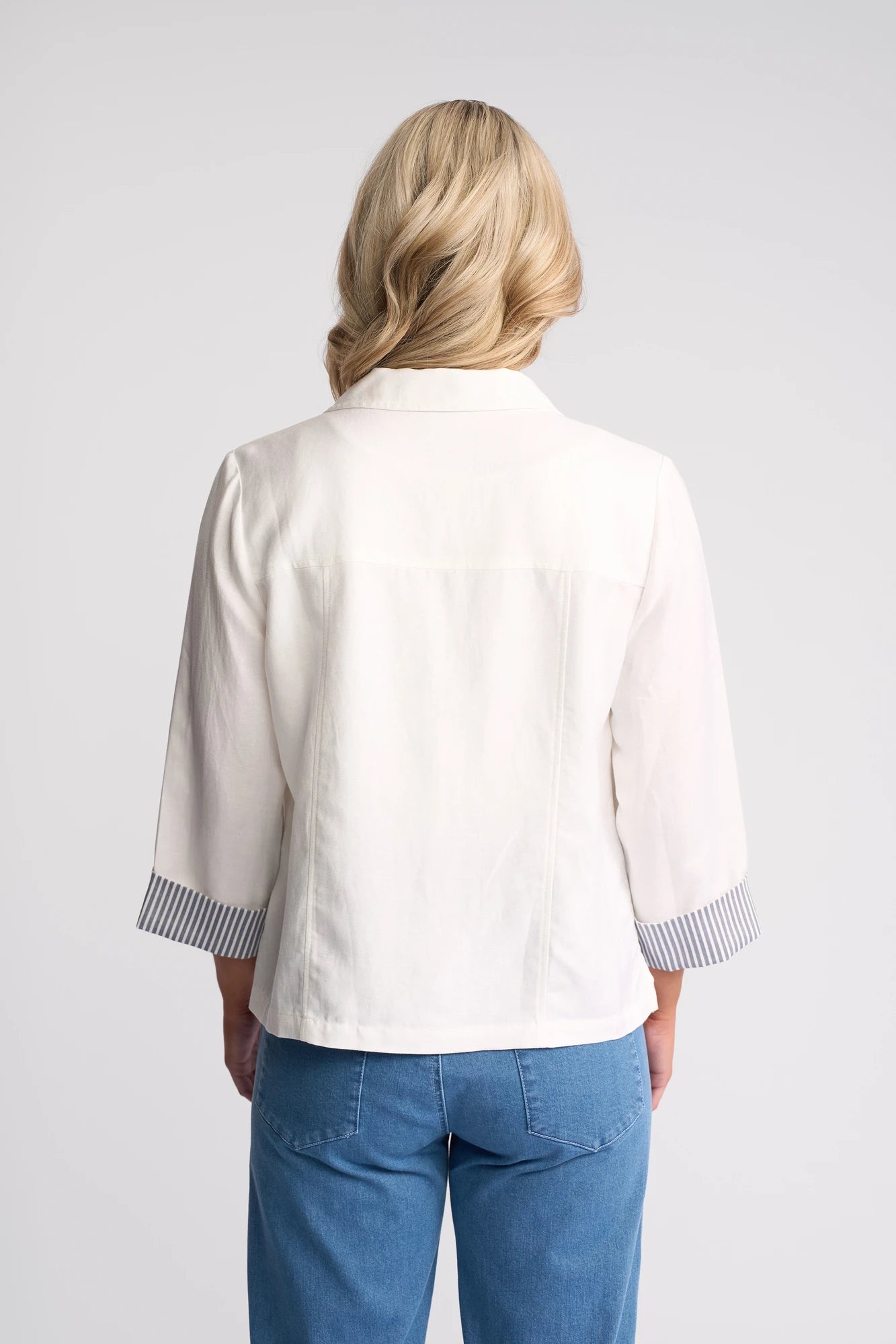 A woman with blonde hair, shown from the back, wears a lightweight Vassalli Honey Jacket in White featuring striped cuffs, paired with blue jeans against a plain light background.