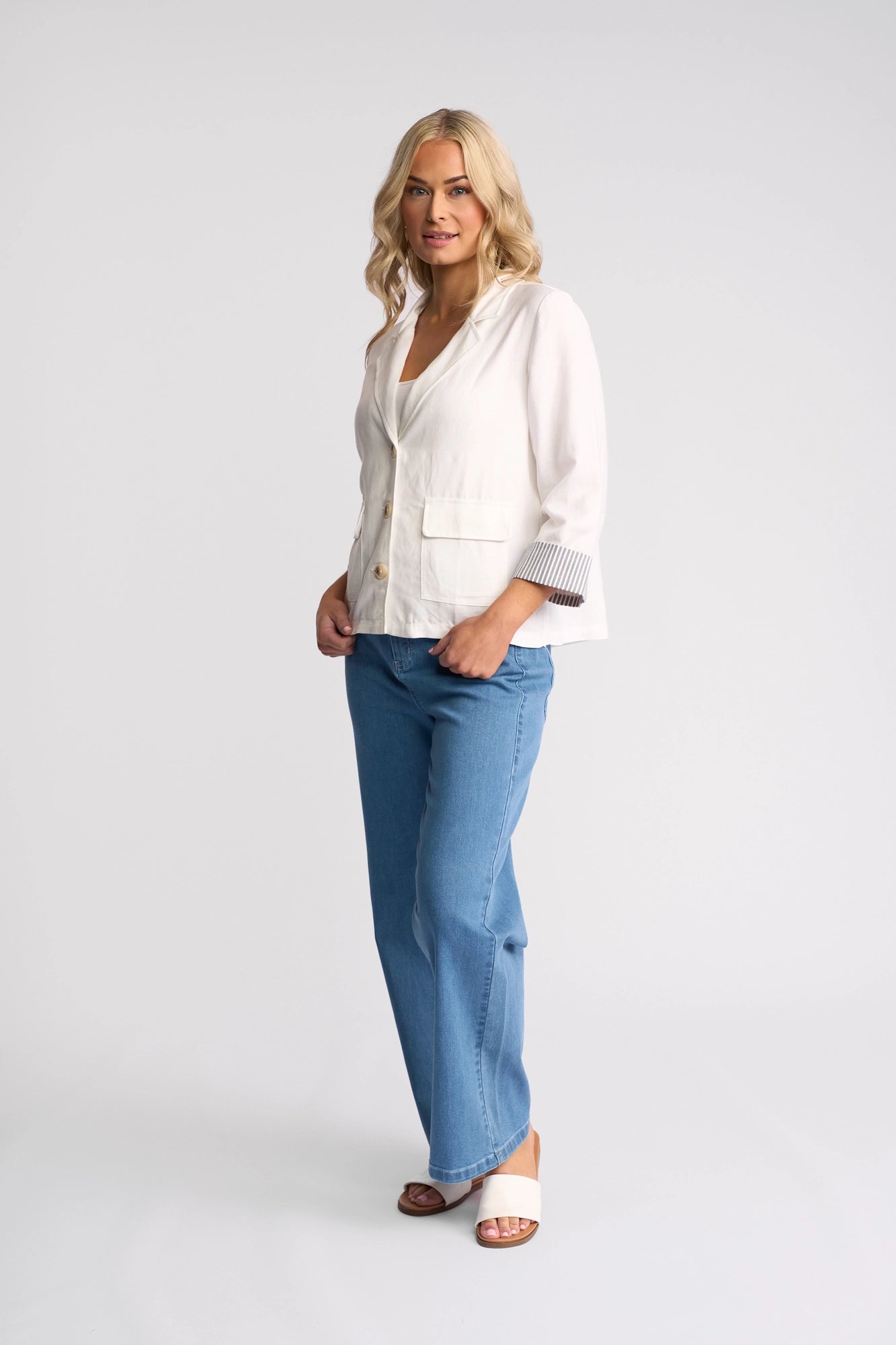 A woman with blonde hair stands in a lightweight linen-viscose Vassalli Honey Jacket in White, paired with blue jeans and white sandals, against a plain light background, looking at the camera.